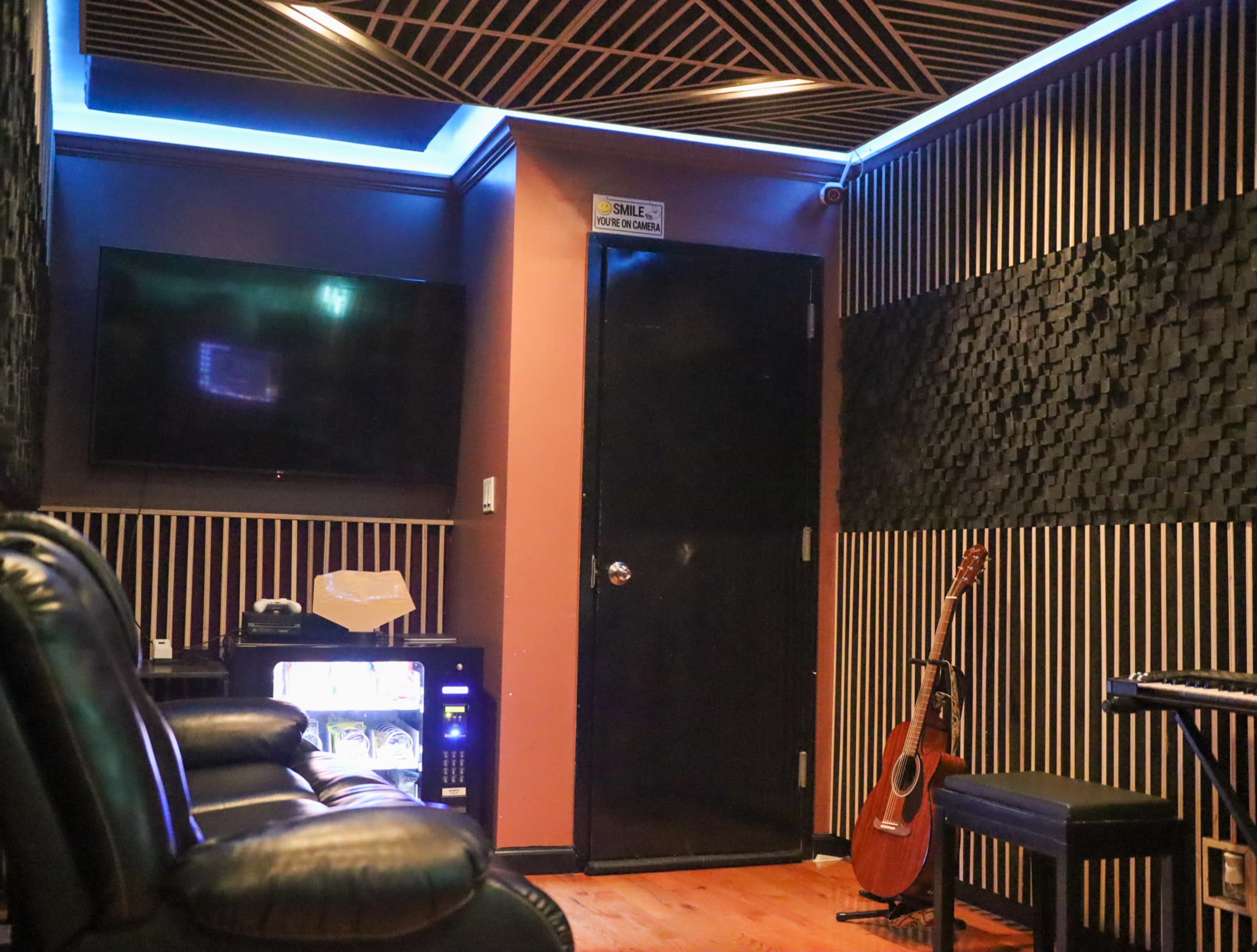 The image shows a cozy room with a television mounted on the wall, a black door, an acoustic guitar leaning against a wall, and soundproof paneling.