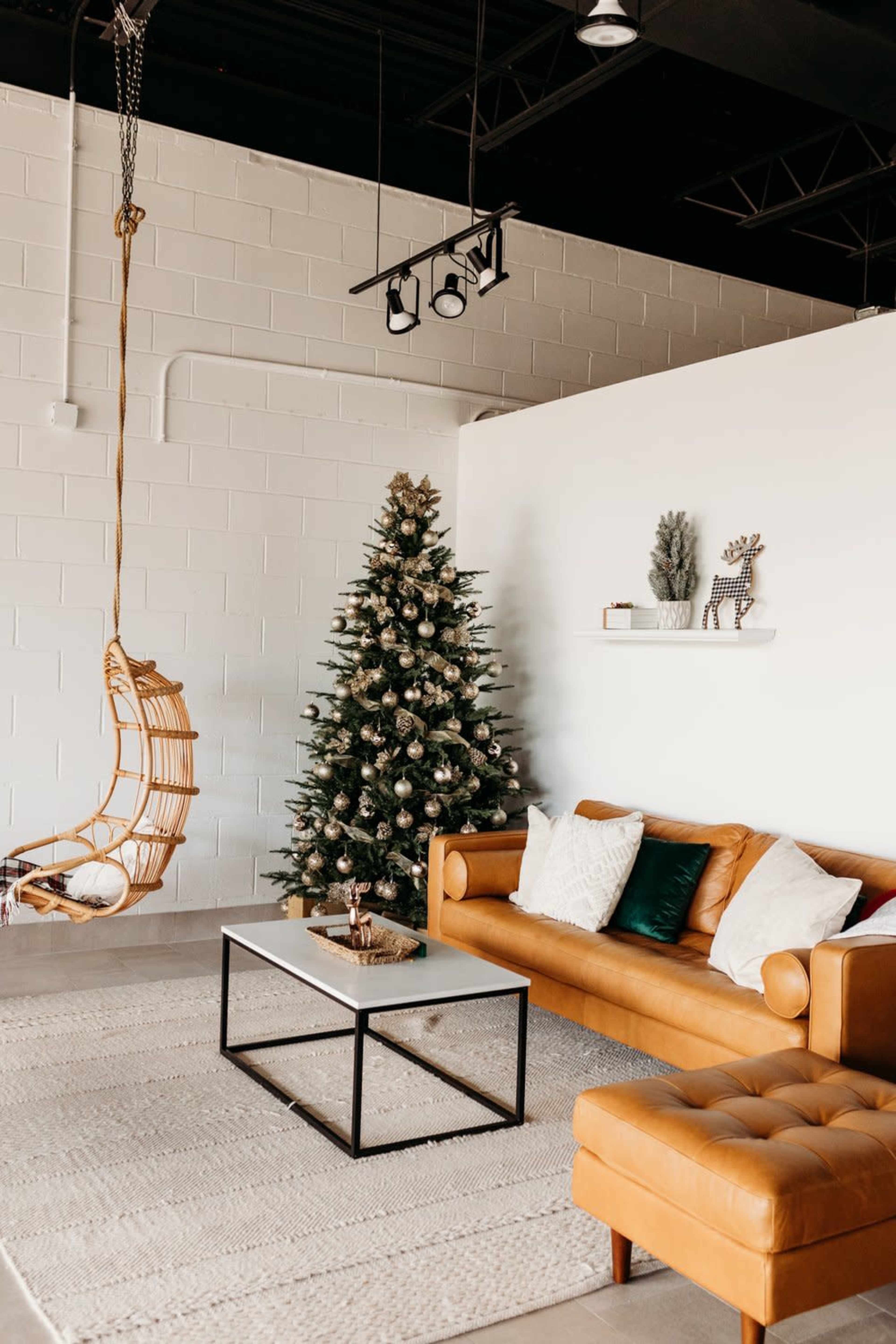 A modern living room features a brown leather sofa, a coffee table, a hanging chair, a decorated Christmas tree, and a neutral-toned rug.