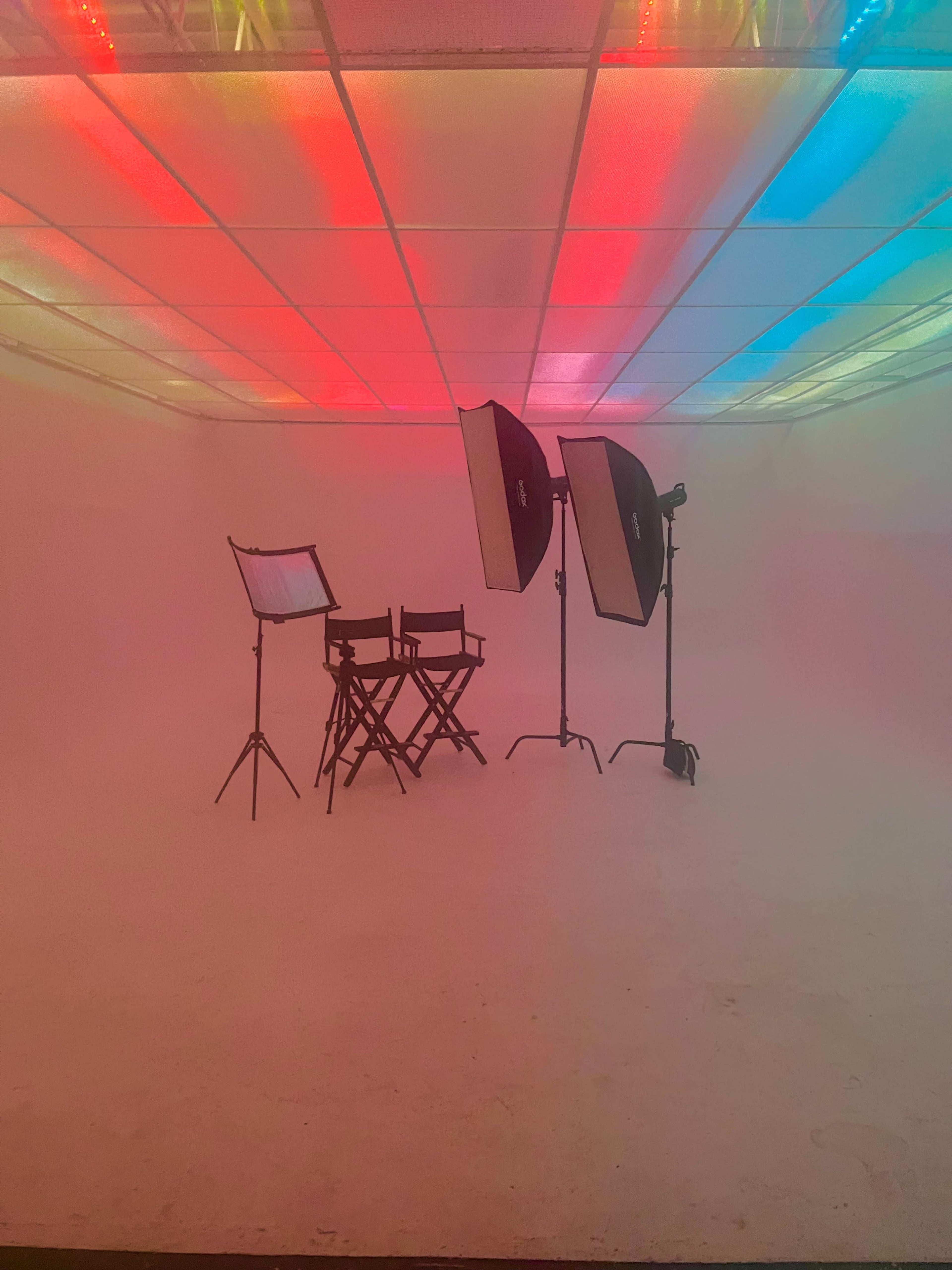 A photography studio is set up with a director's chair, a script holder, and two large lights, all against a softly lit, multicolored background.