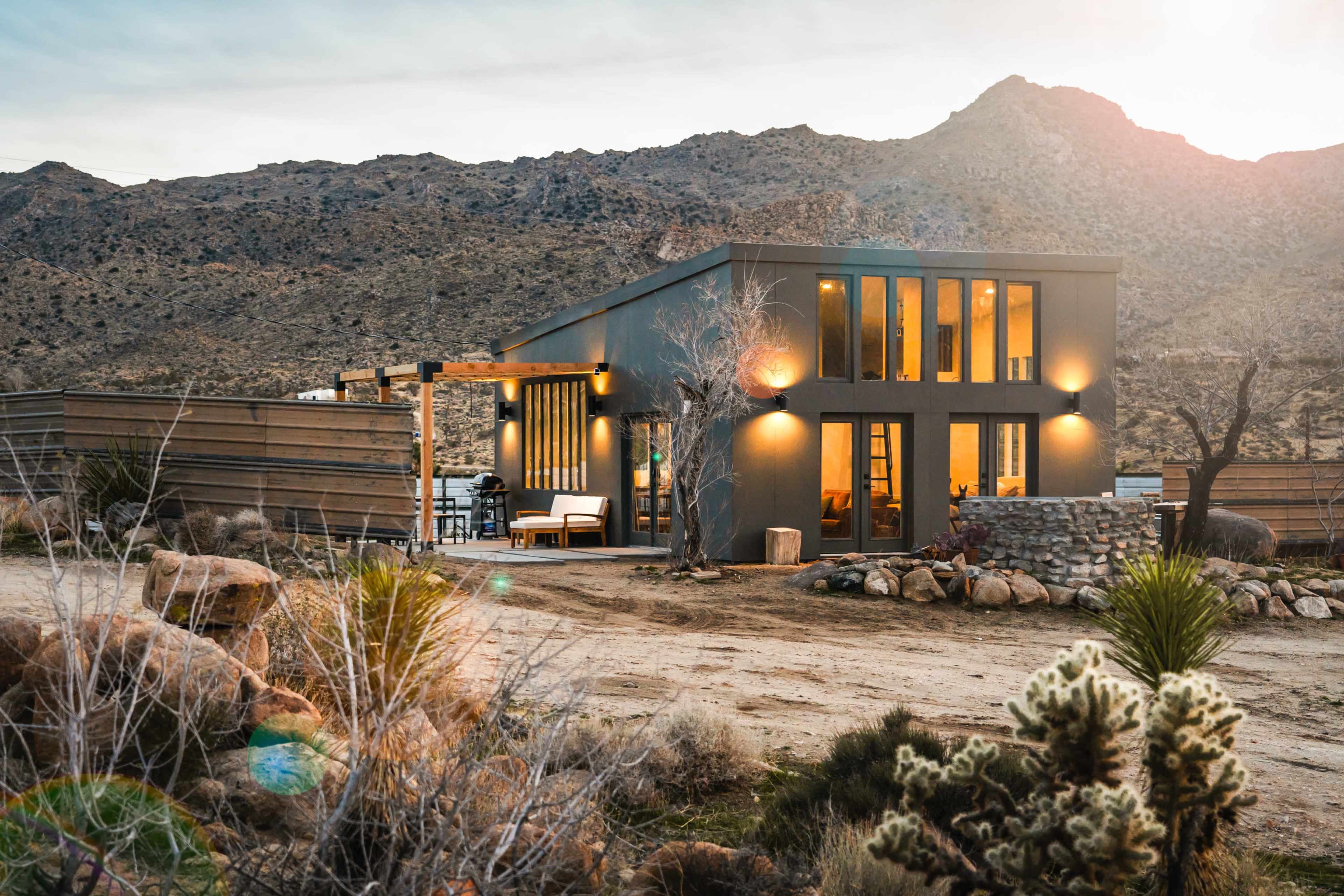 A modern house with large windows is situated in a rugged mountainous landscape, surrounded by desert vegetation.