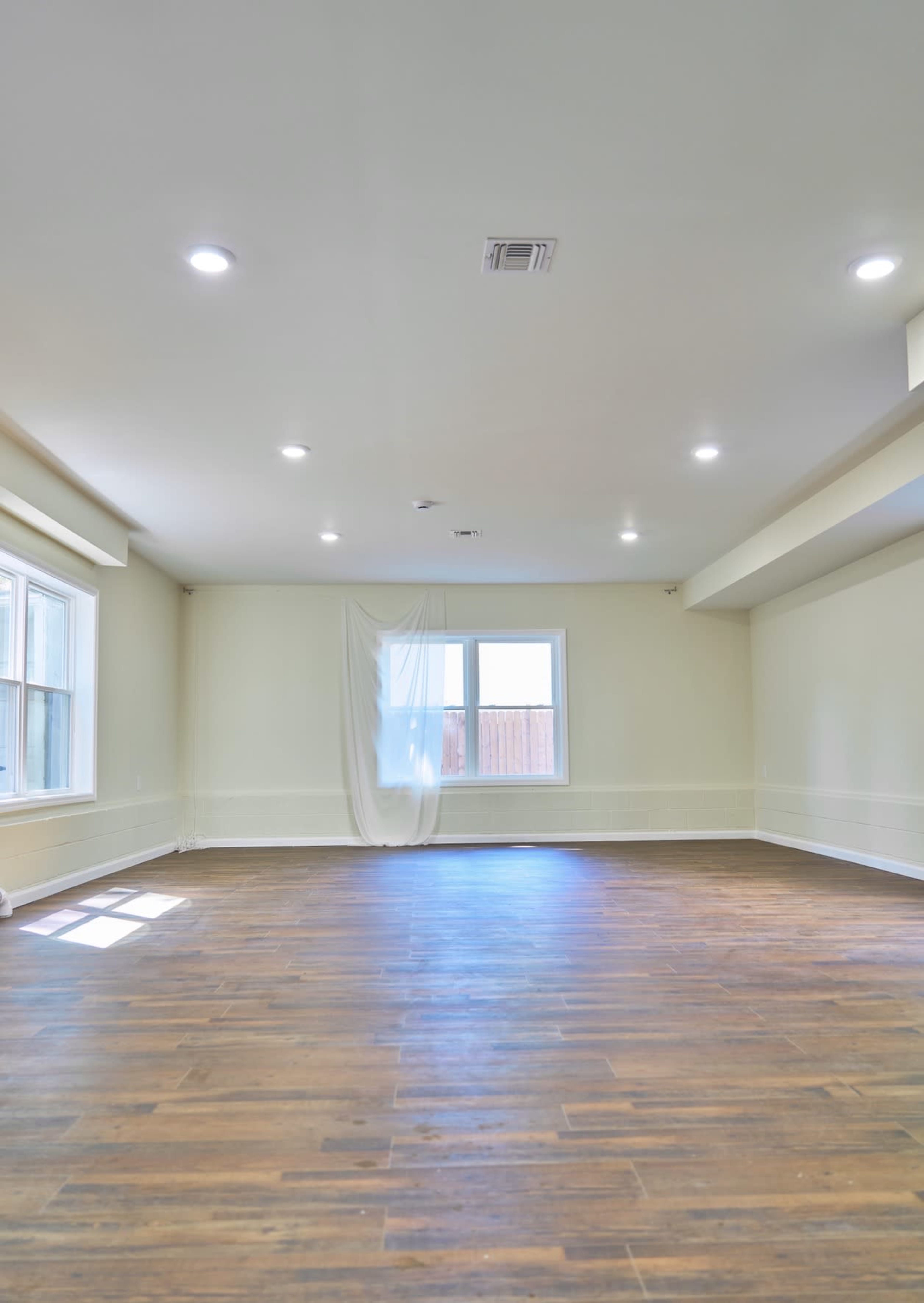 The image shows an empty room with a wooden floor, two windows on one wall, and a white curtain hanging in the center.