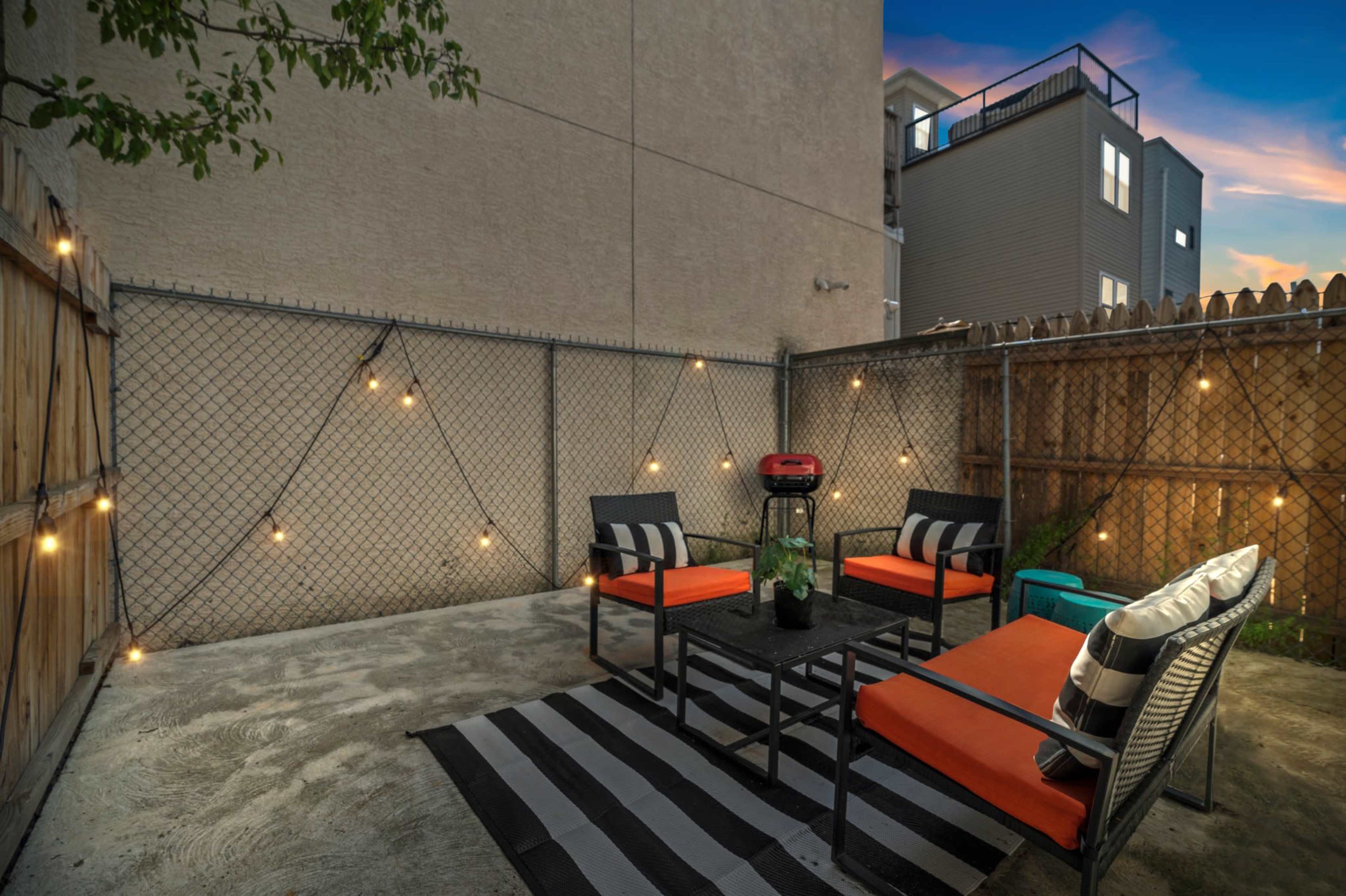 A small outdoor seating area with black and orange furniture, a small grill, and string lights along a chain-link fence.