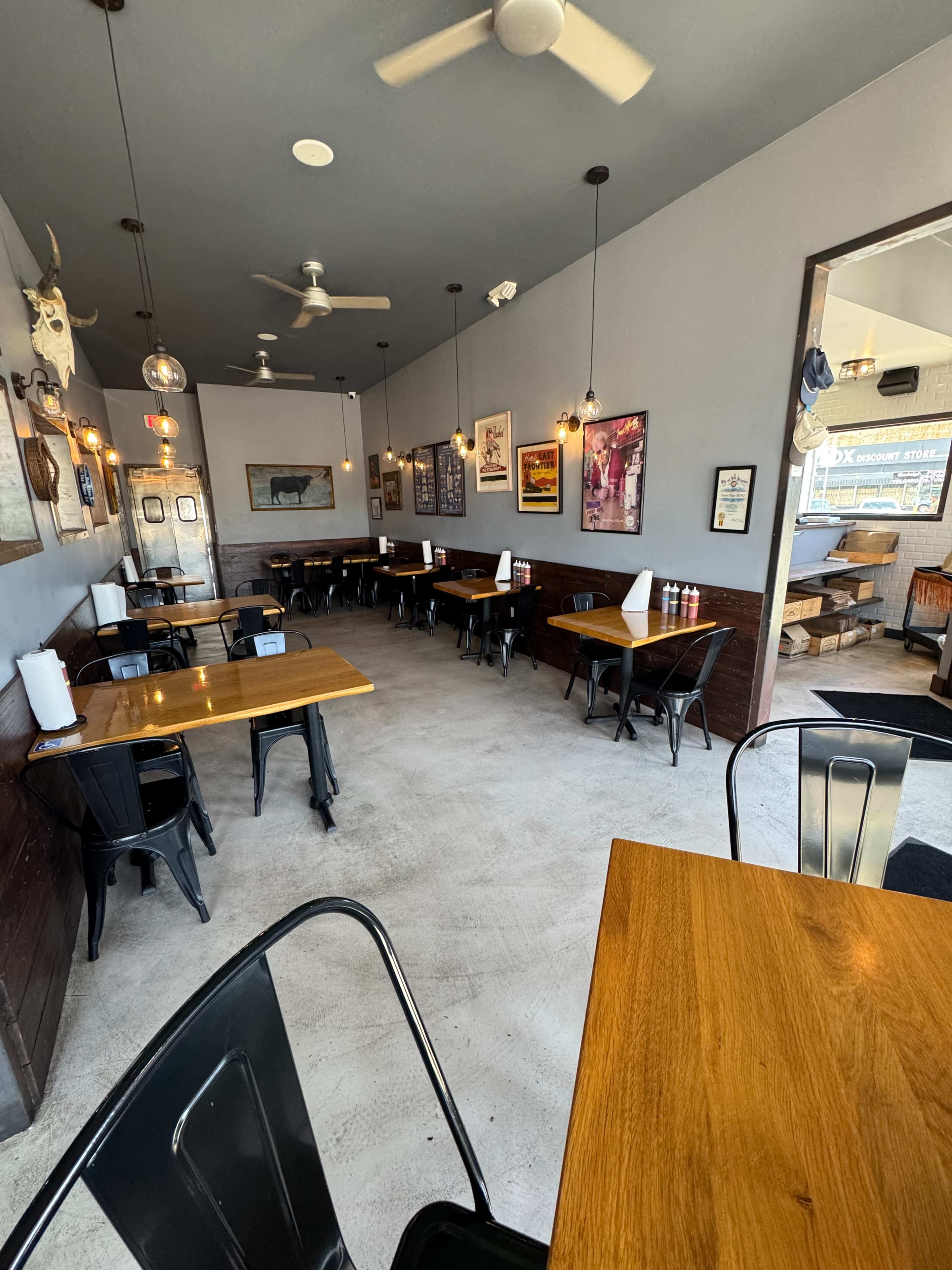 The image shows an empty restaurant interior with wooden tables, black chairs, and wall-mounted decorations under soft lighting.