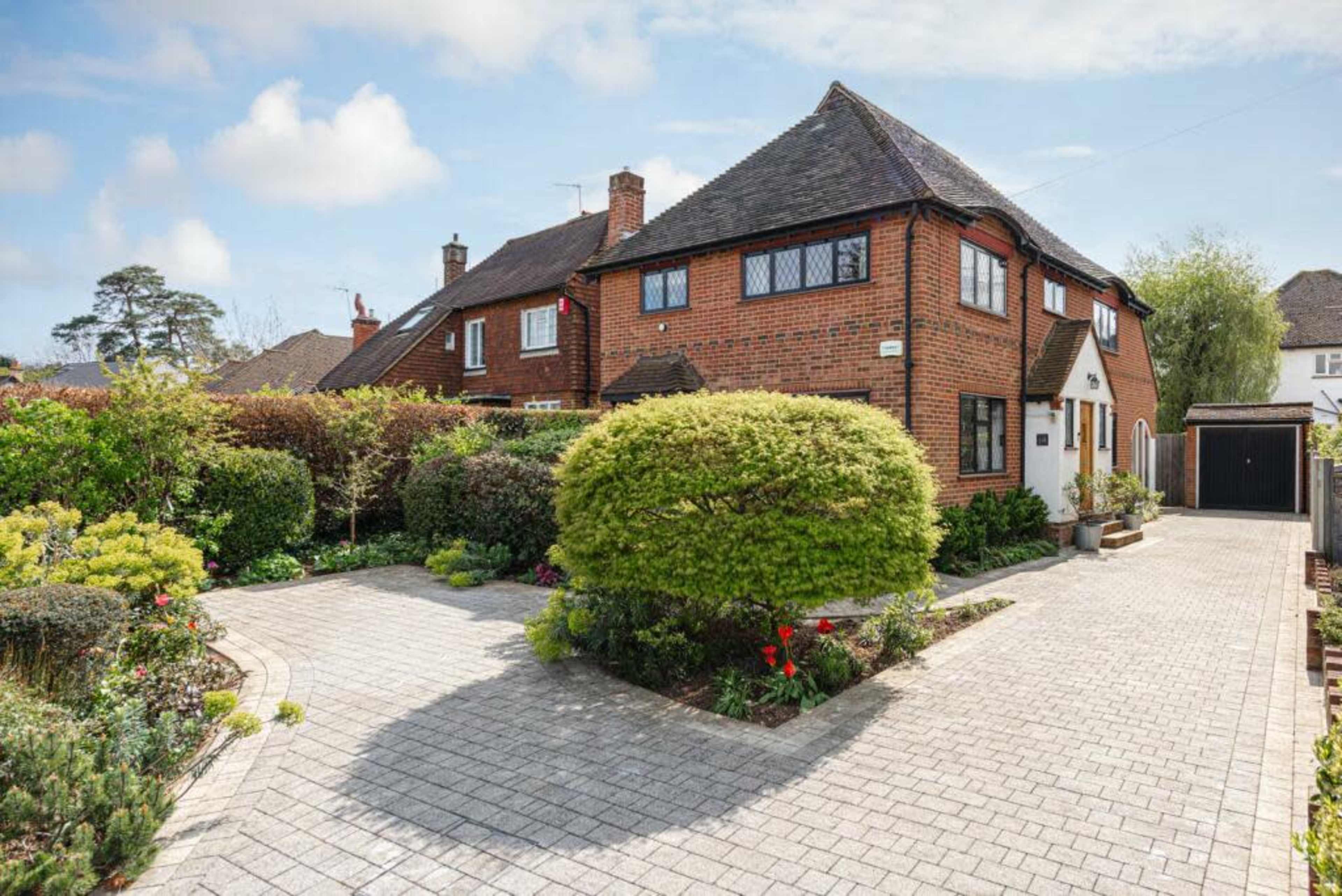 A red-brick house with a driveway and neatly landscaped gardens is surrounded by greenery and neighboring homes.