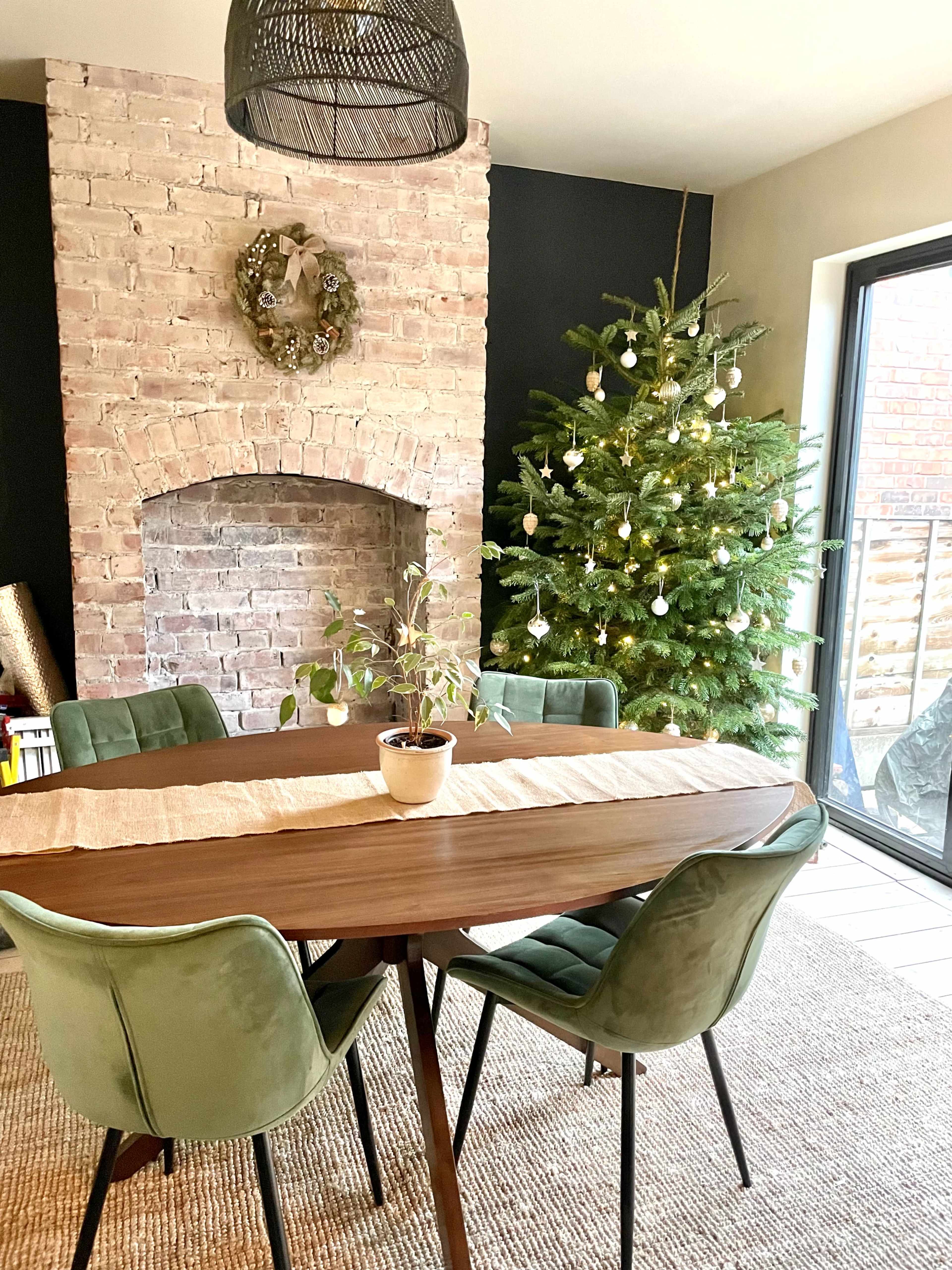 A dining area features a wooden table surrounded by green chairs, with a brick fireplace and a decorated Christmas tree in the background.