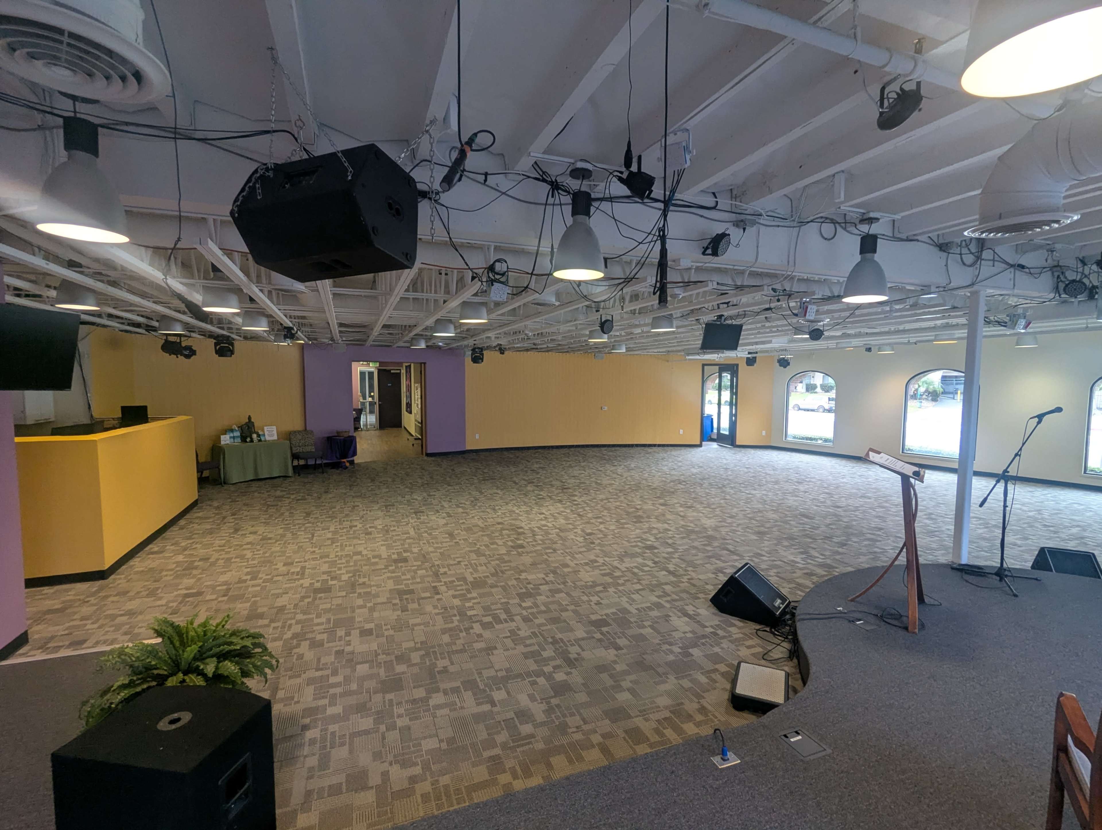 The image shows a spacious, empty room with a carpeted floor, overhead lighting, and a small stage area equipped with a microphone and stand.