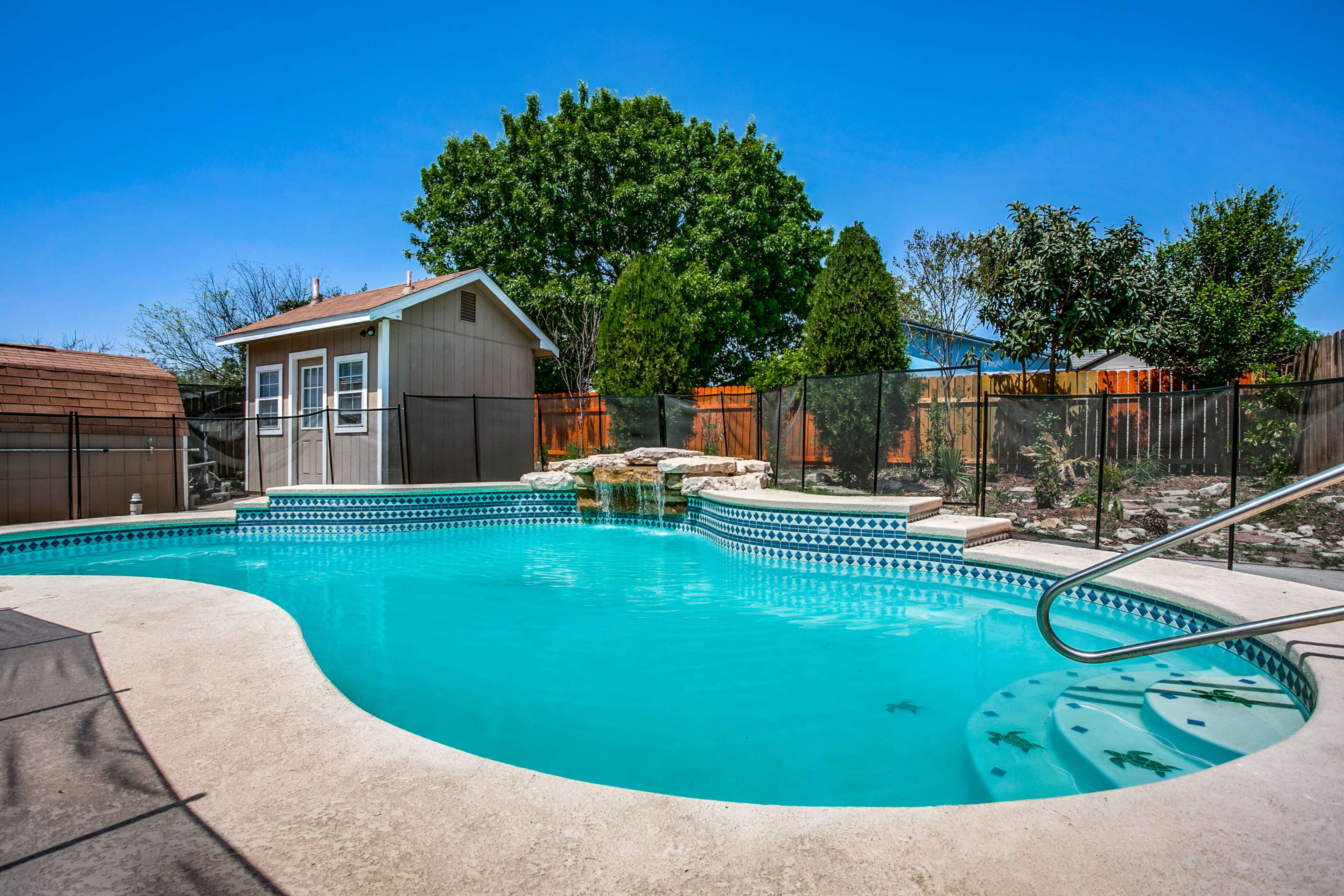 A backyard swimming pool with a waterfall feature, surrounded by a fence and a small shed in the background.