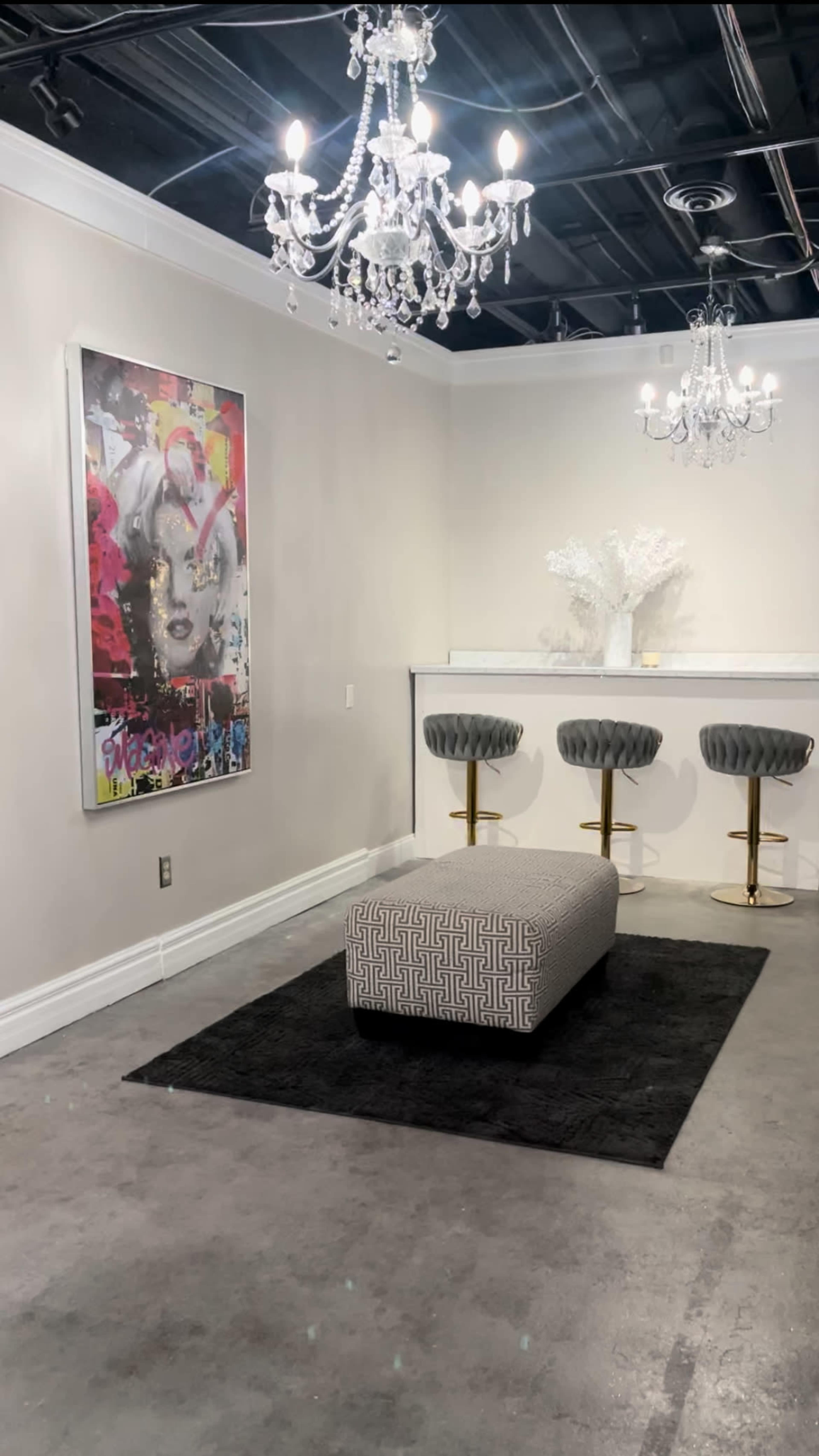 The image shows a modern interior space featuring a bar area with three gray stools, a patterned ottoman in the center, and a large artwork on the wall under decorative chandeliers.