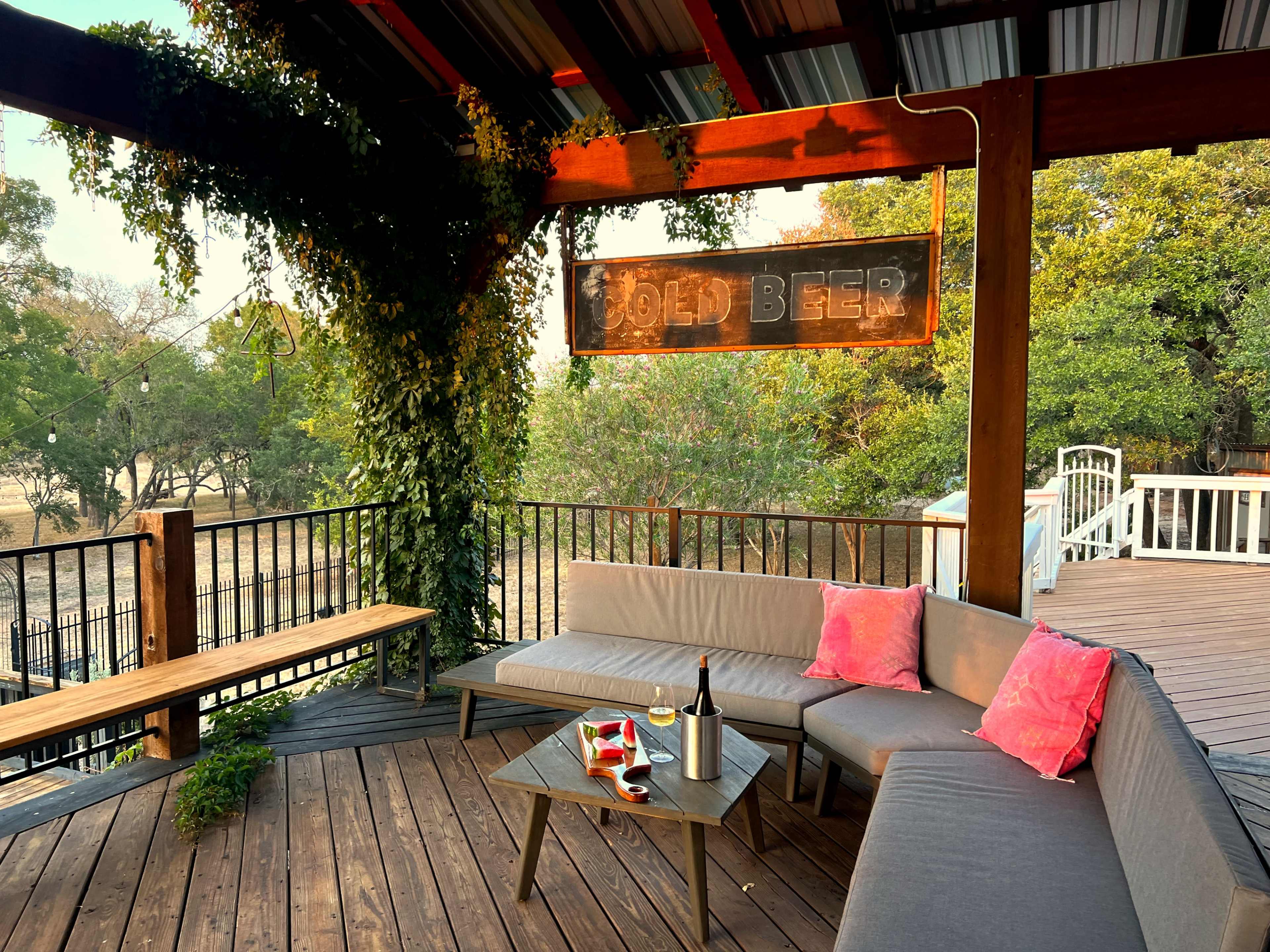 A covered outdoor patio features a lounging area with gray sectional seating, bright pink pillows, a small table, and a sign that reads "Cold Beer."