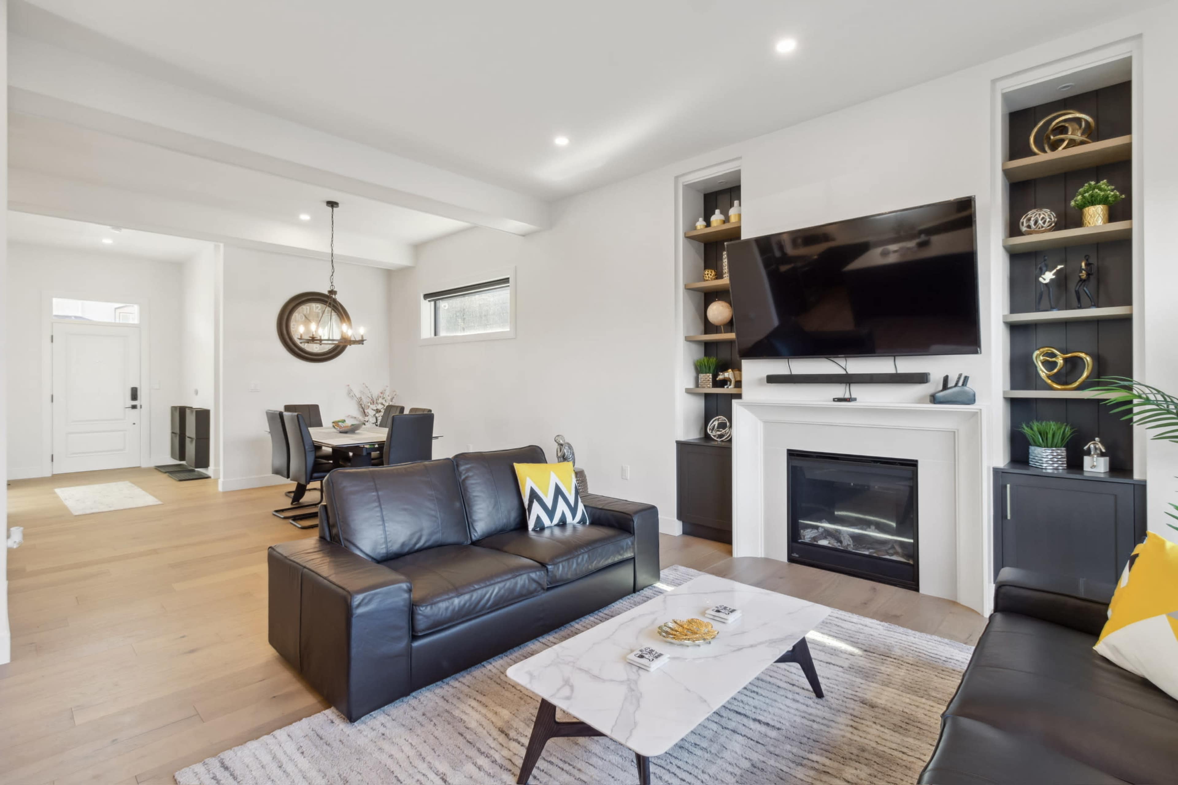 A modern living room features black leather sofas, a marble coffee table, built-in shelves with decorative items, and a wall-mounted television above a fireplace.