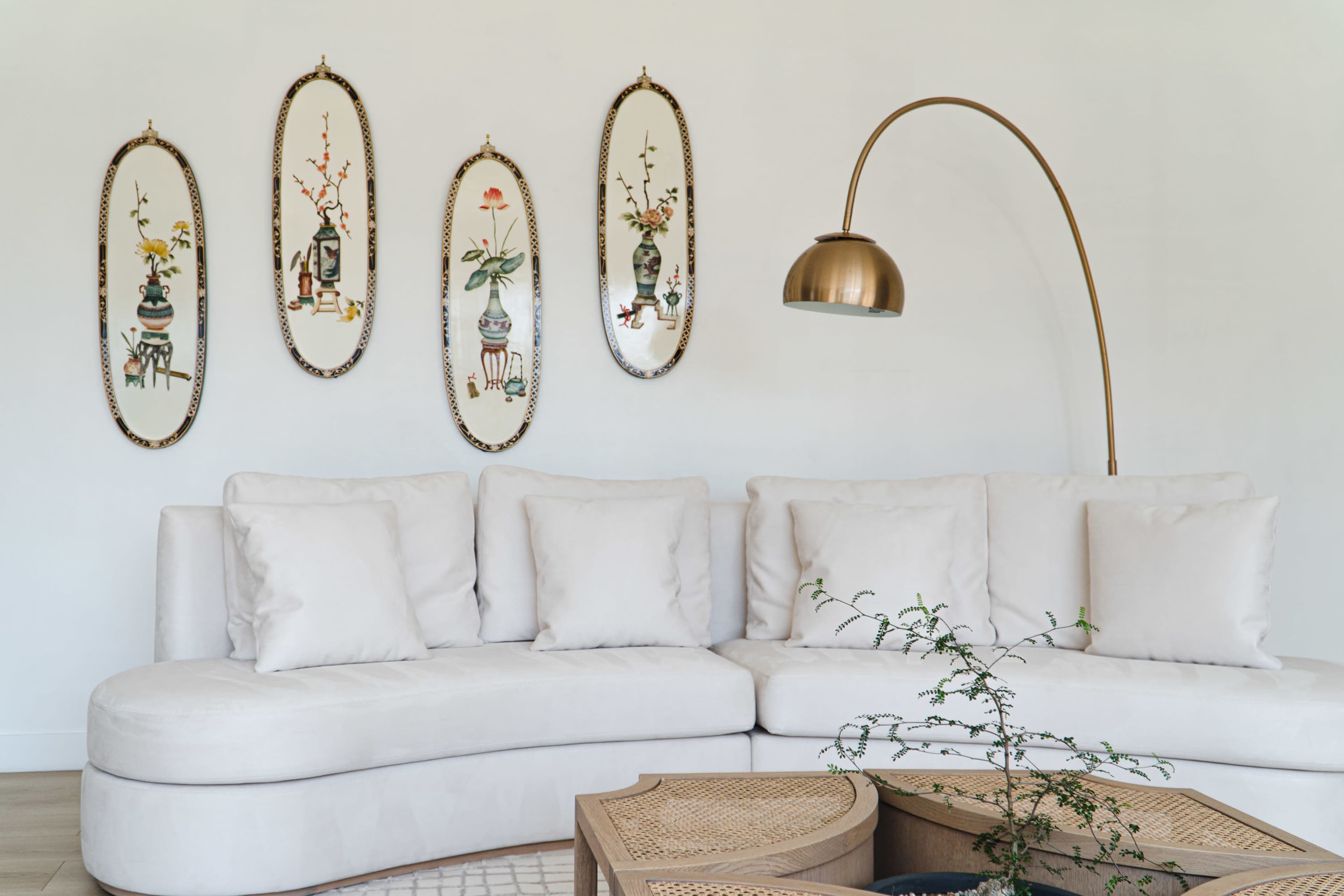 The image shows a light-colored sectional sofa with several plush cushions, facing a decorative wall featuring five framed botanical illustrations.