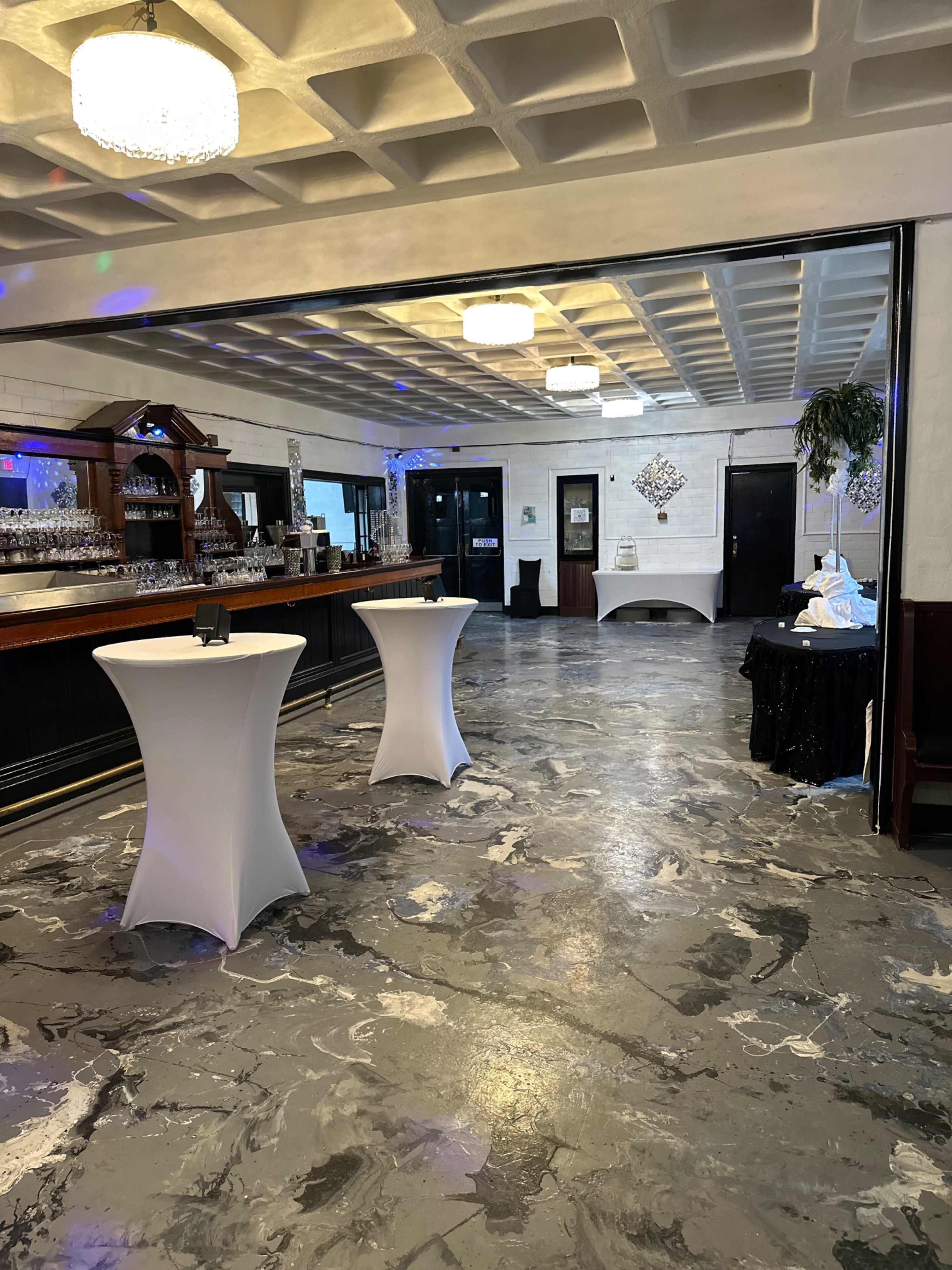 The image shows an event space with a bar in the background, two white cocktail tables in the foreground, and a polished concrete floor.