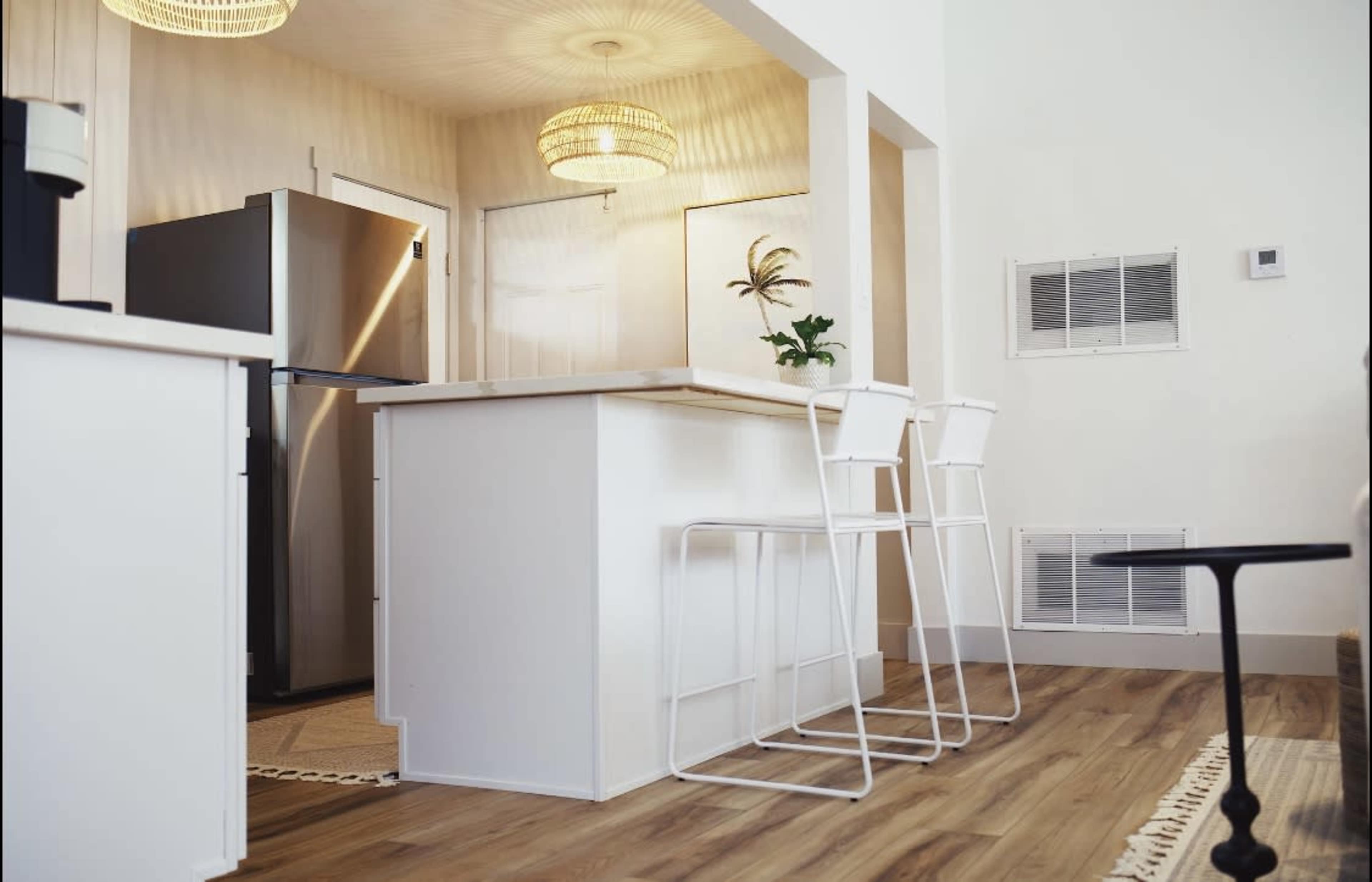 The image shows a modern kitchen with a central island, bar stools, stainless steel refrigerator, and warm, ambient lighting.