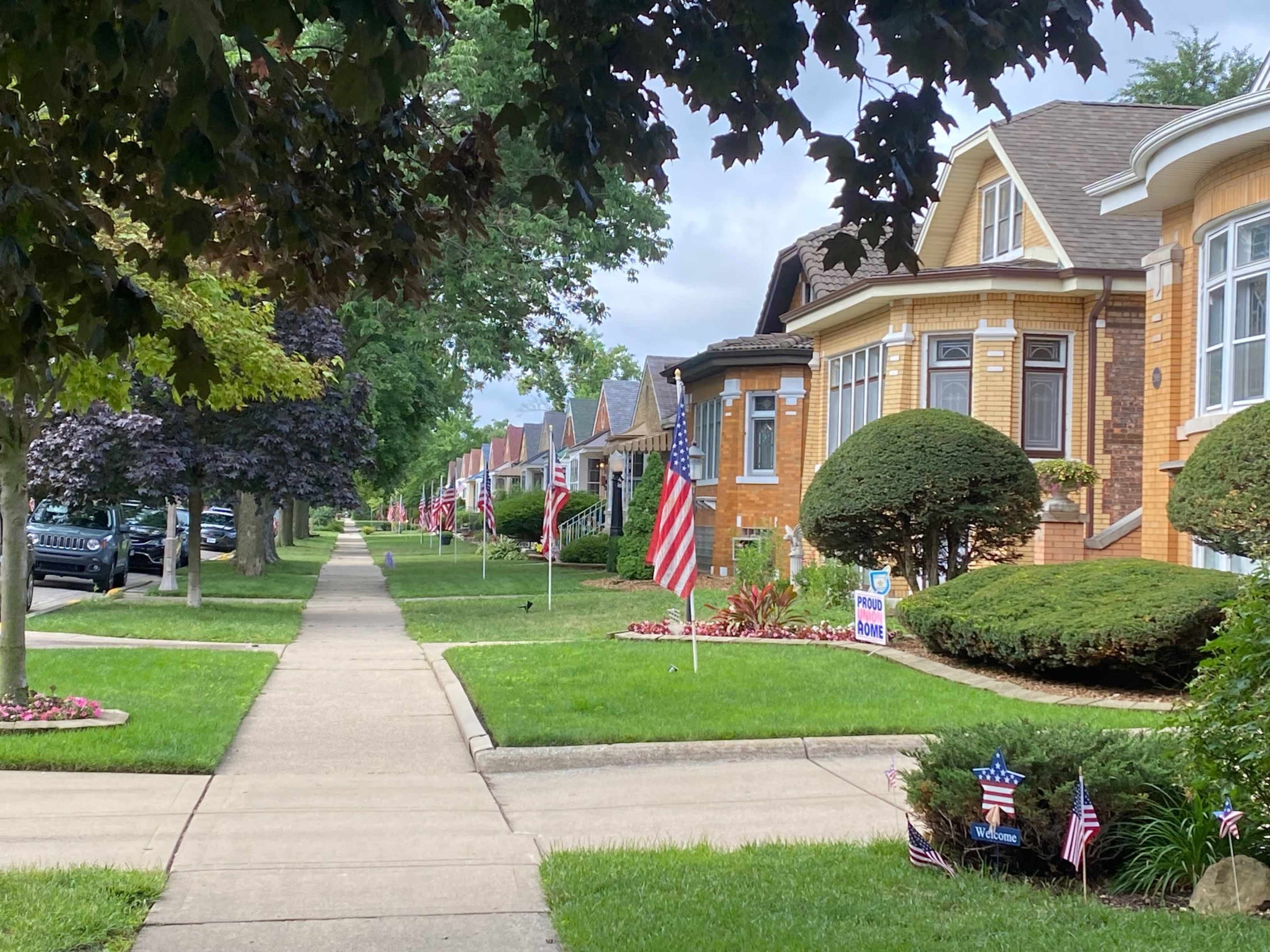 A suburban street lined with houses, each displaying American flags along the grassy lawns and sidewalks.