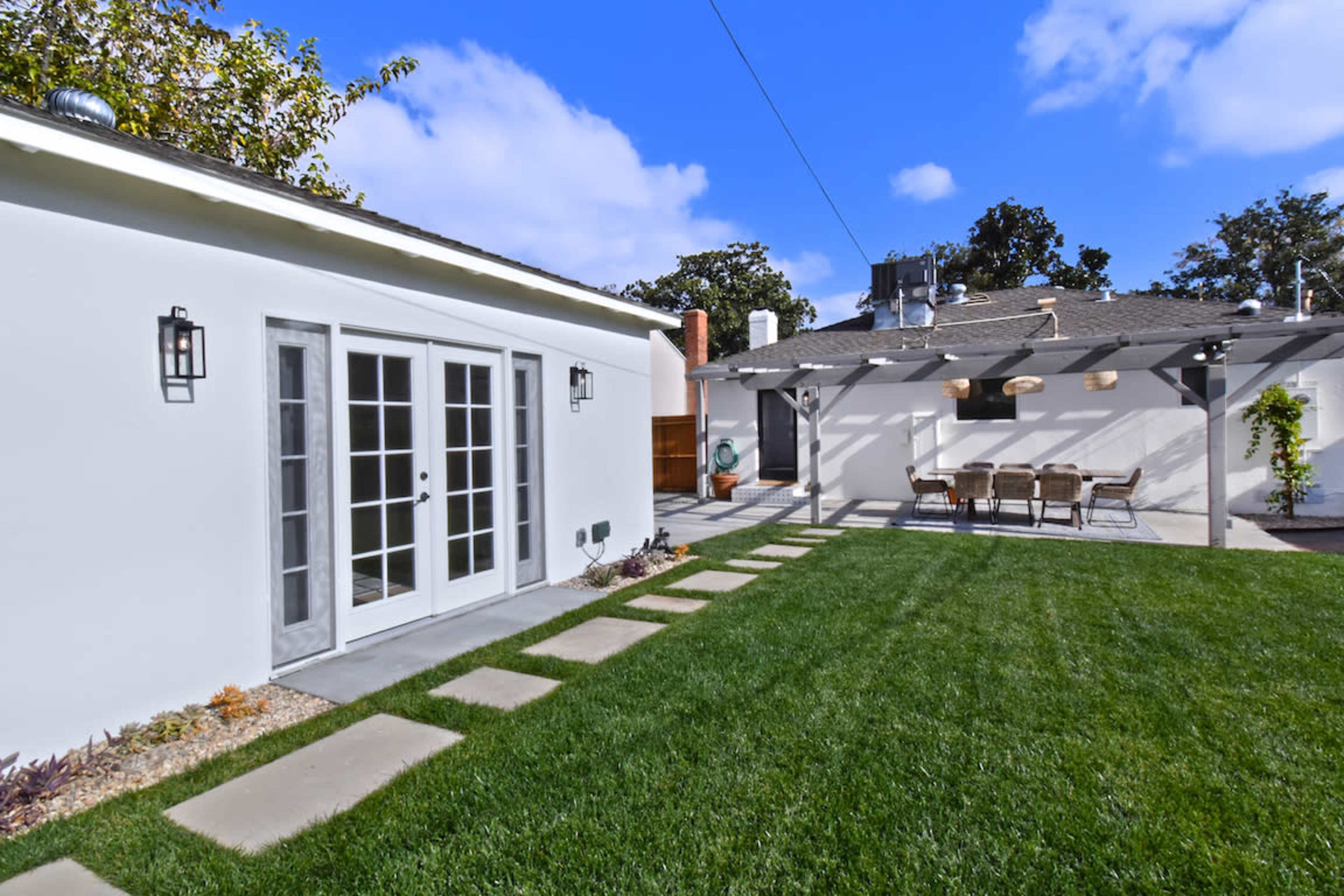 The image shows a backyard with a green lawn, a pathway made of stone pavers, and two white buildings, one with French doors and the other with a patio area.