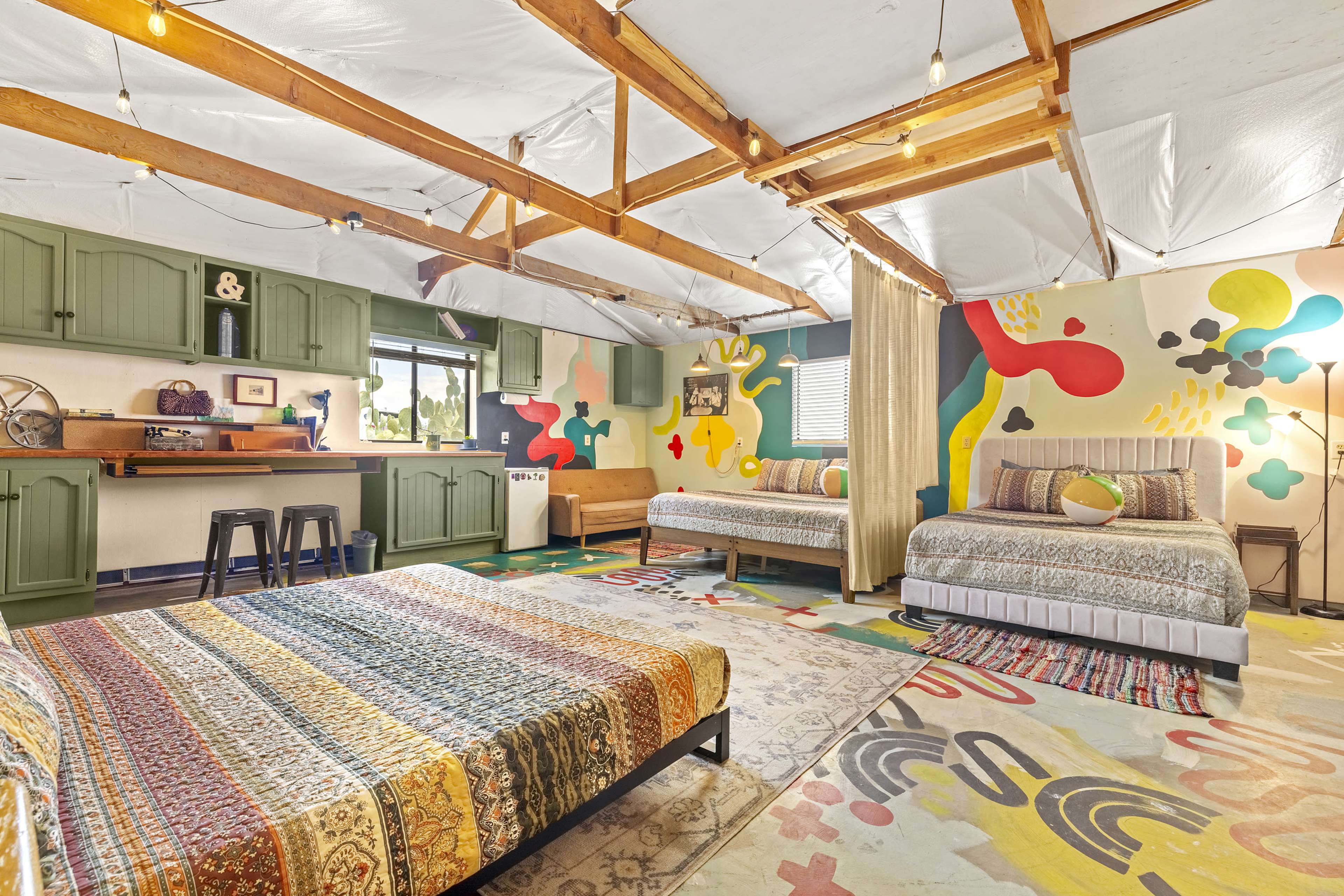 The image shows a colorful bedroom featuring two beds, a kitchenette area, and vibrant wall murals.