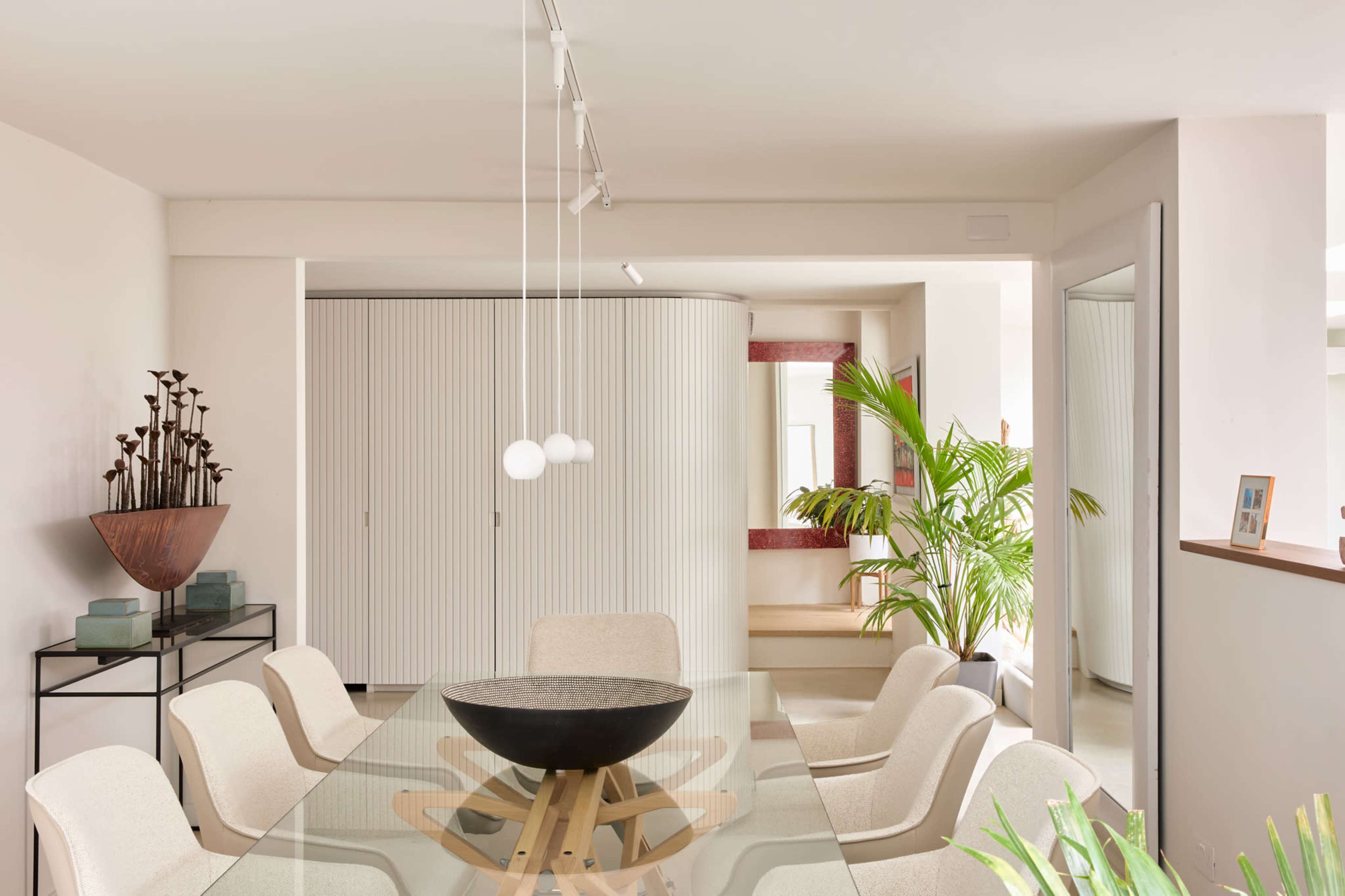 The image shows a modern dining area featuring a glass table surrounded by upholstered chairs, with a decorative bowl at the center and plants in the background.