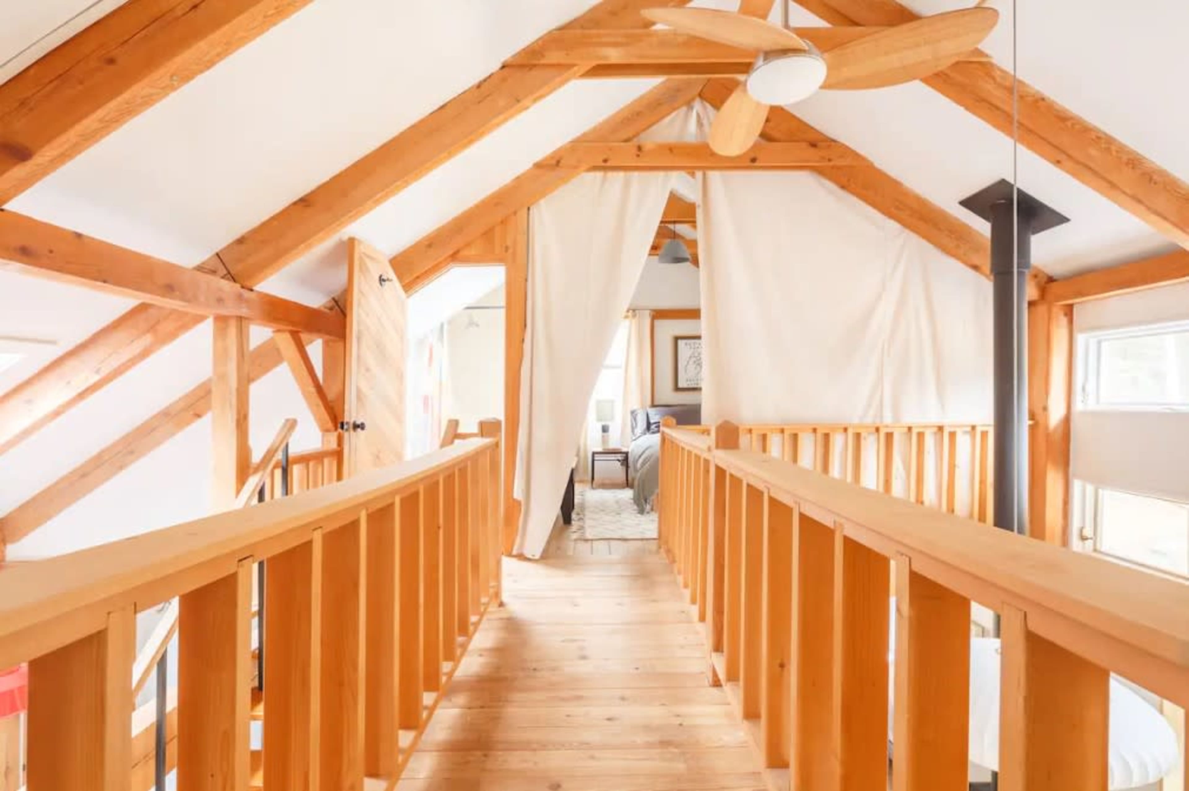 The image shows a wooden loft area with exposed beams, featuring a railing along a hallway that leads to a room with light-colored walls and furnishings.