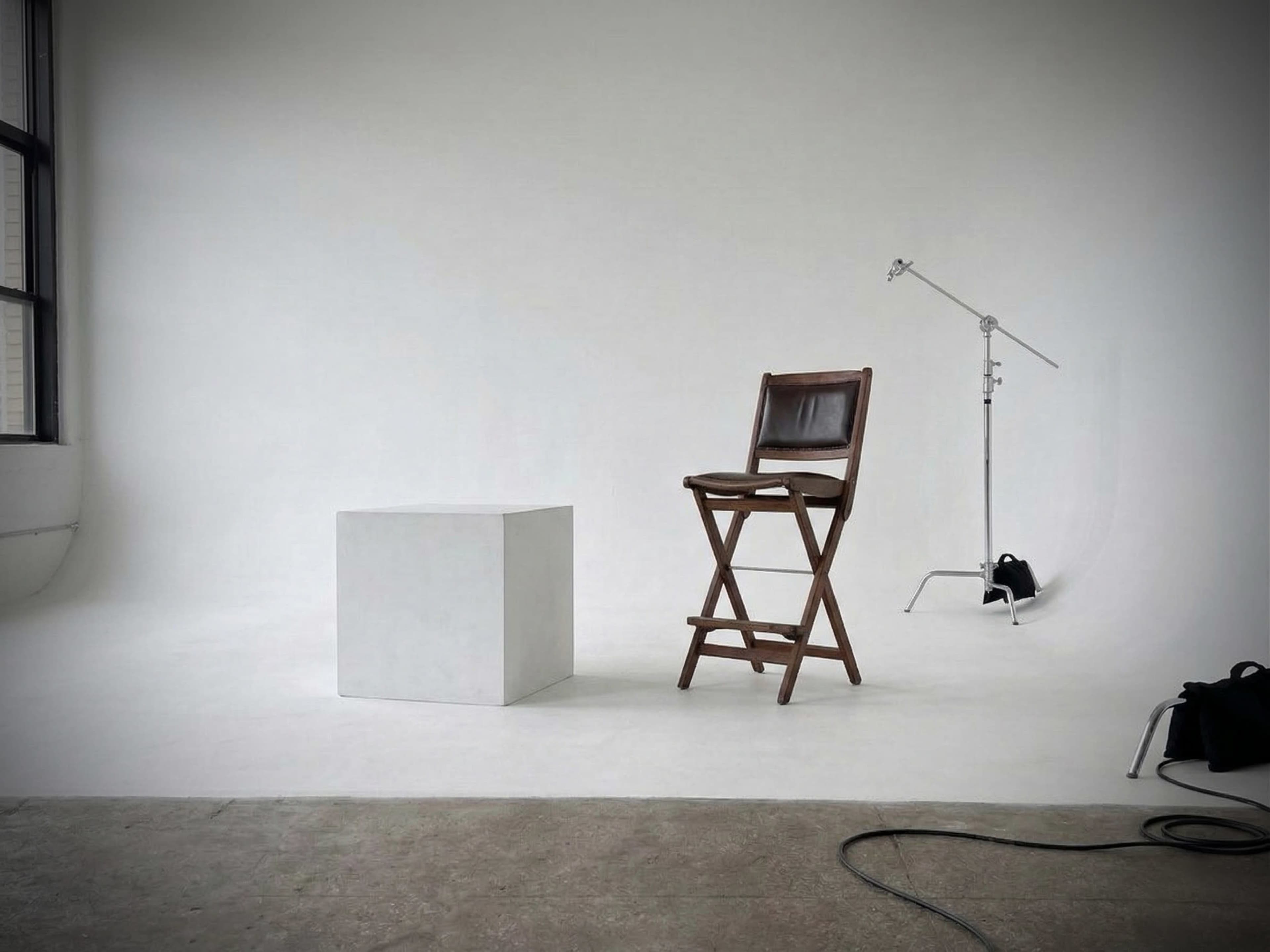 The image shows a minimalist photo studio with a wooden chair placed next to a white cube, equipped with a lighting stand and a black bag in the background.