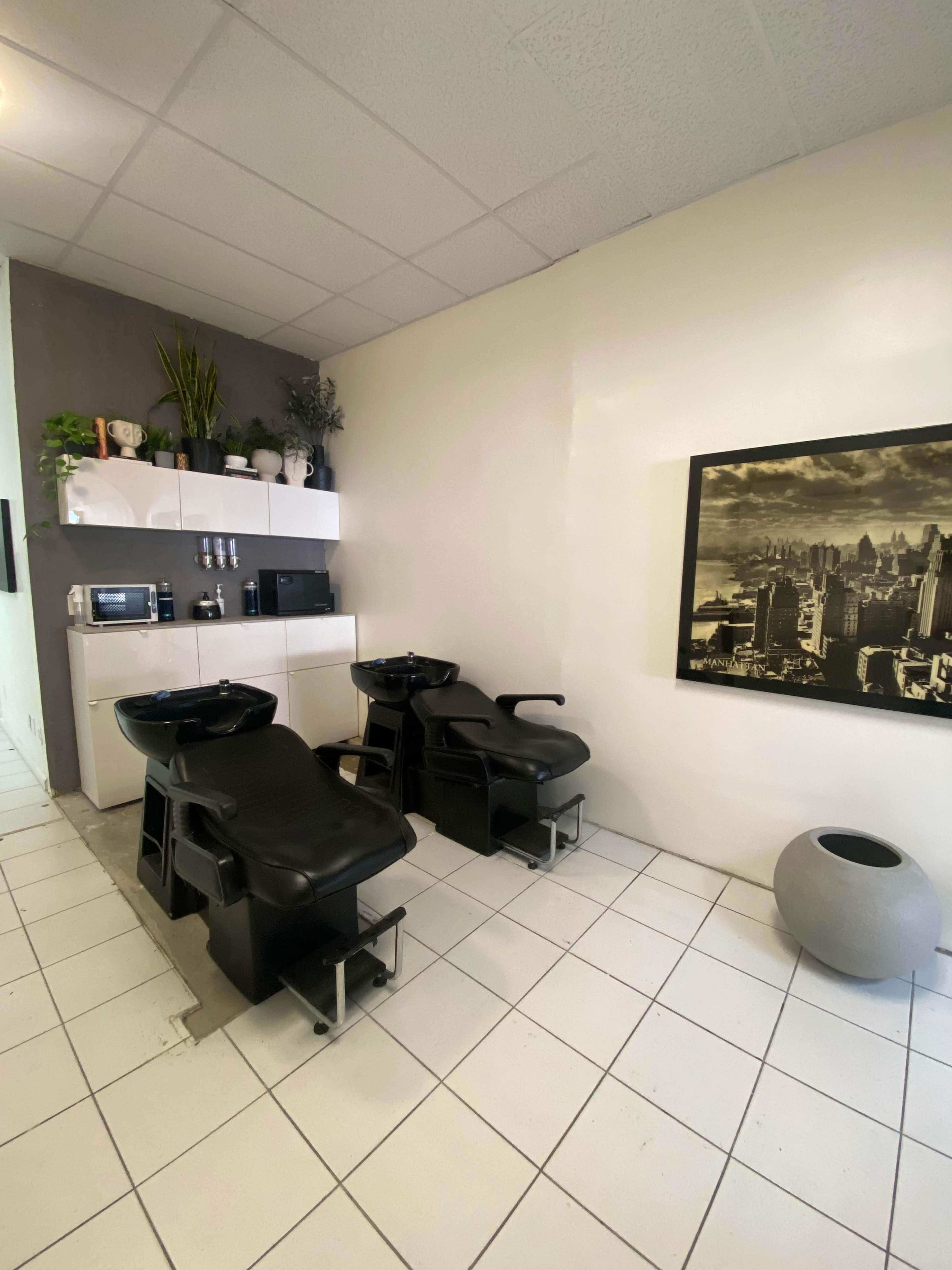 A salon area with two black shampoo chairs, a white shelf with plants and appliances, and a large framed black-and-white photo on the wall.