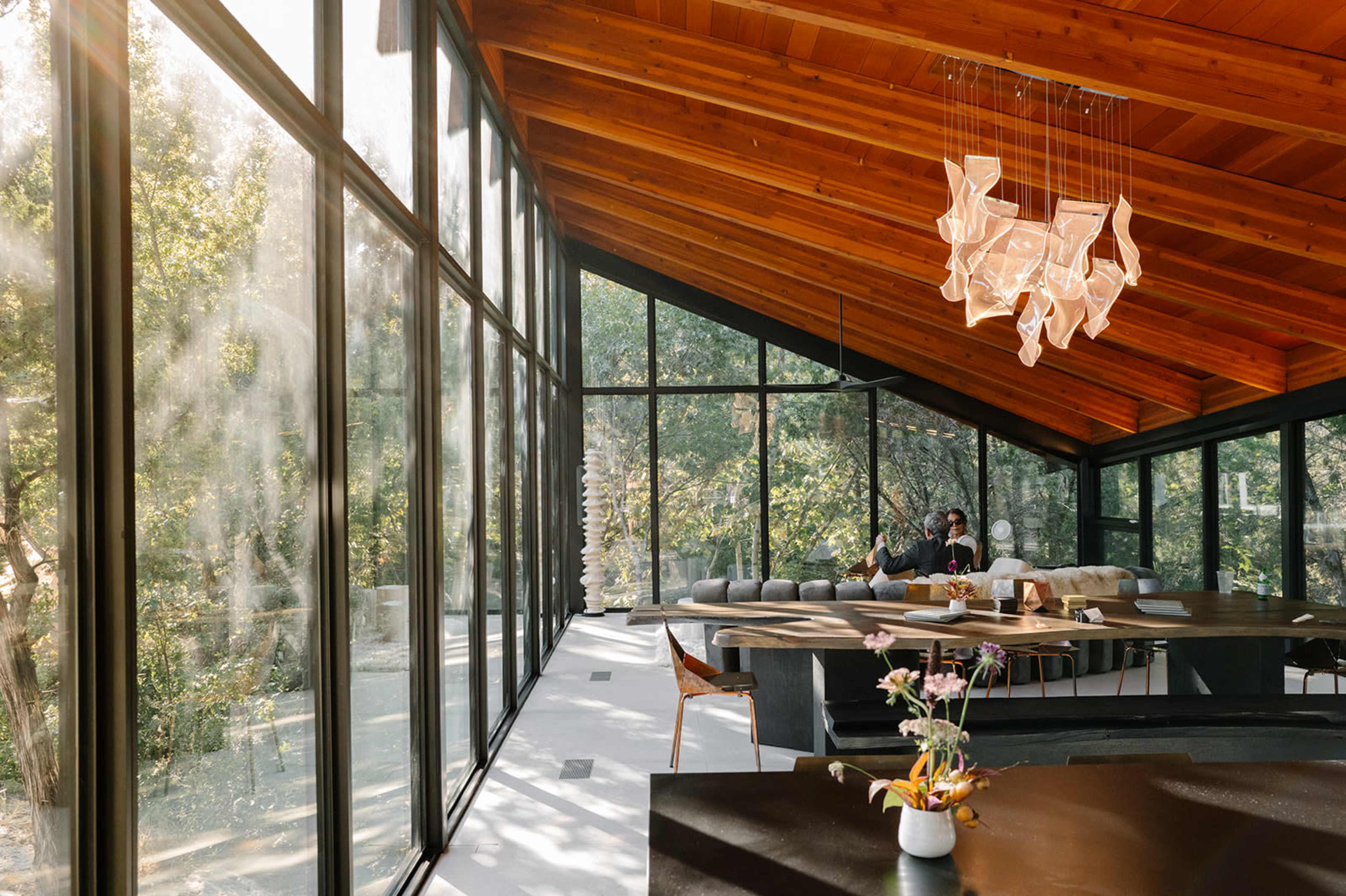 A modern interior features large windows with a wooden ceiling, showcasing a dining area and a view of greenery outside.