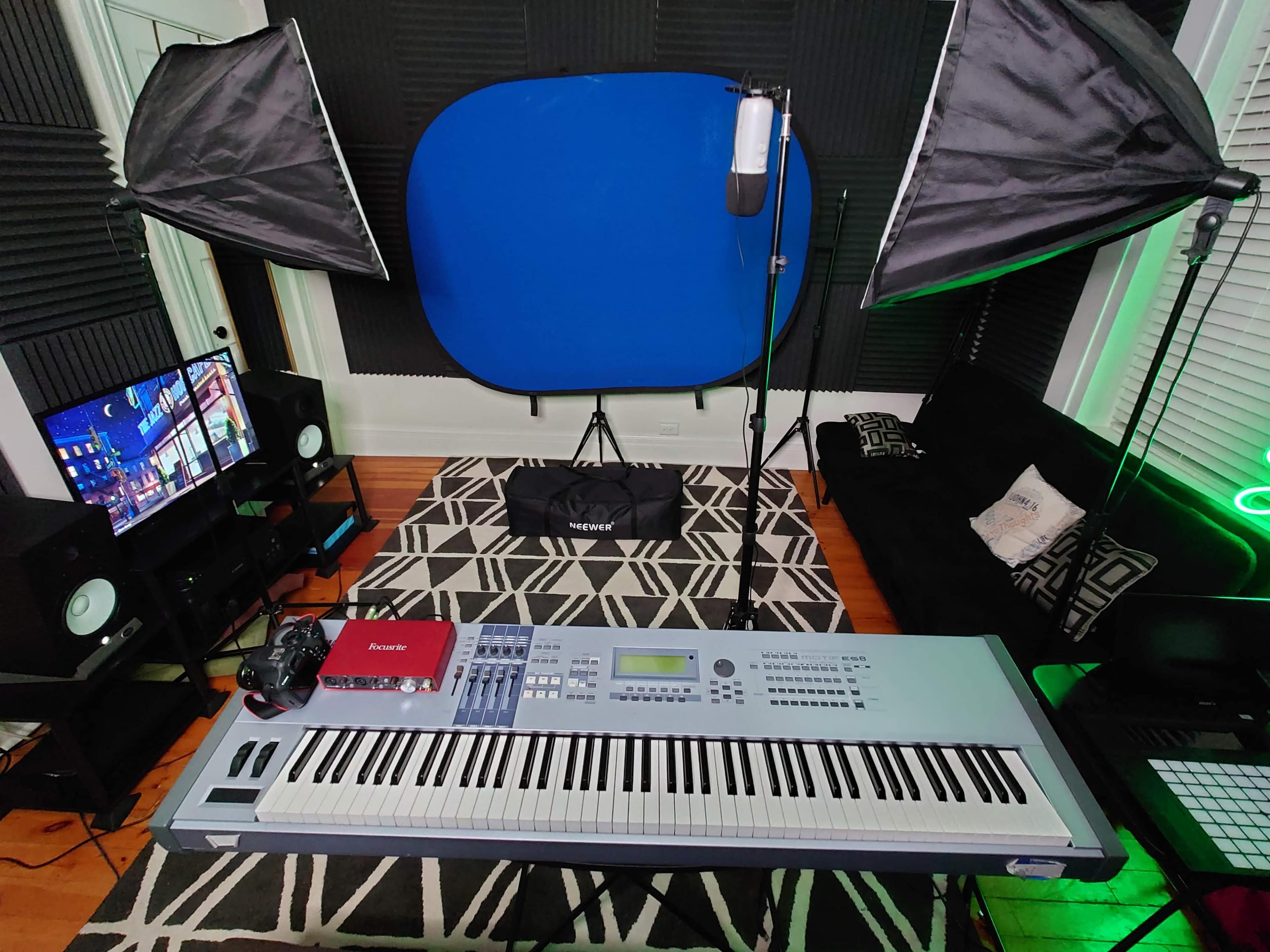 A music production setup with a keyboard, a microphone, lighting equipment, a monitor, and a laptop arranged in a room with acoustic panels and a blue backdrop.