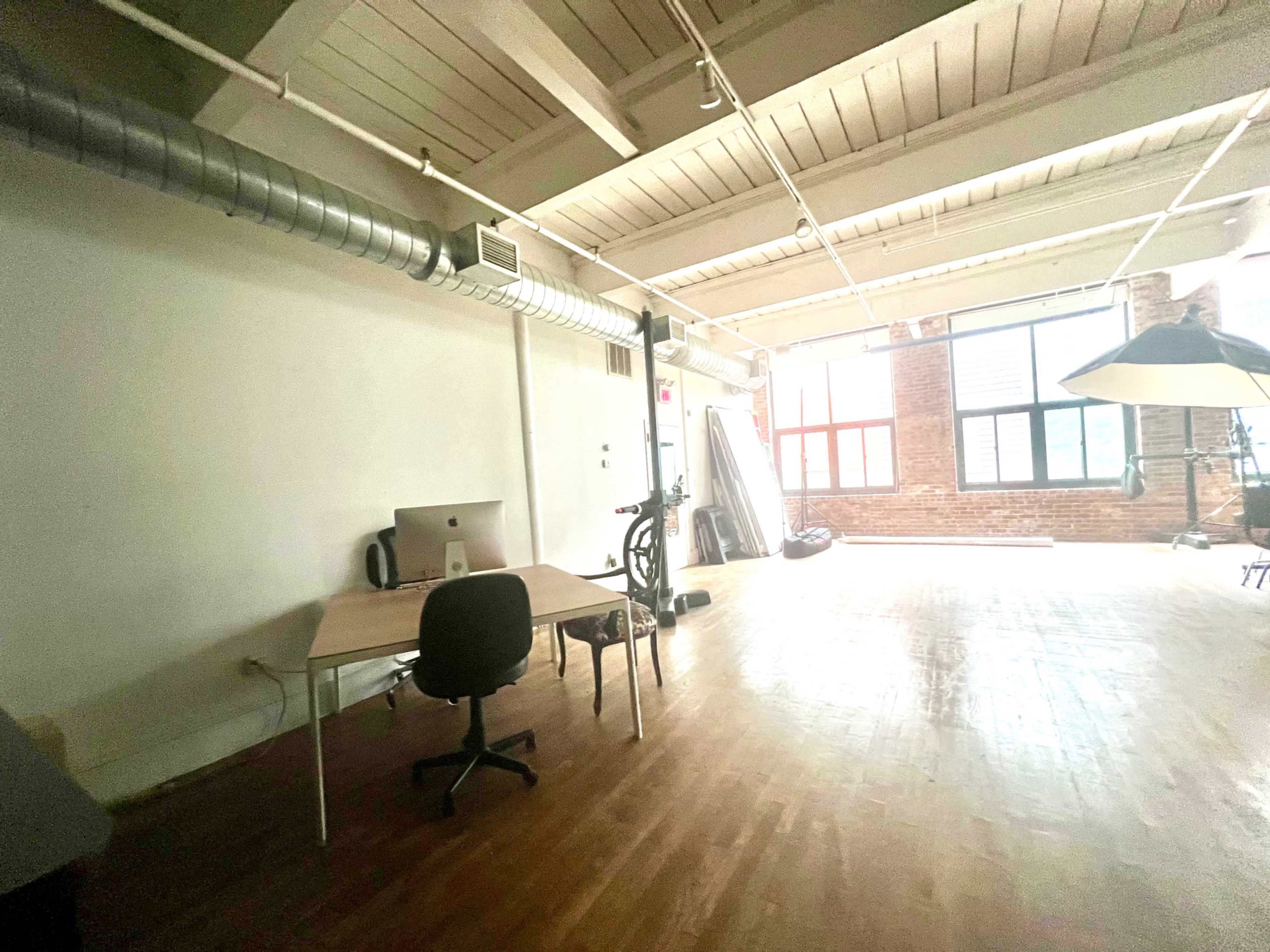 A spacious room with wooden flooring, a desk and chair on one side, and large windows allowing natural light to fill the space.