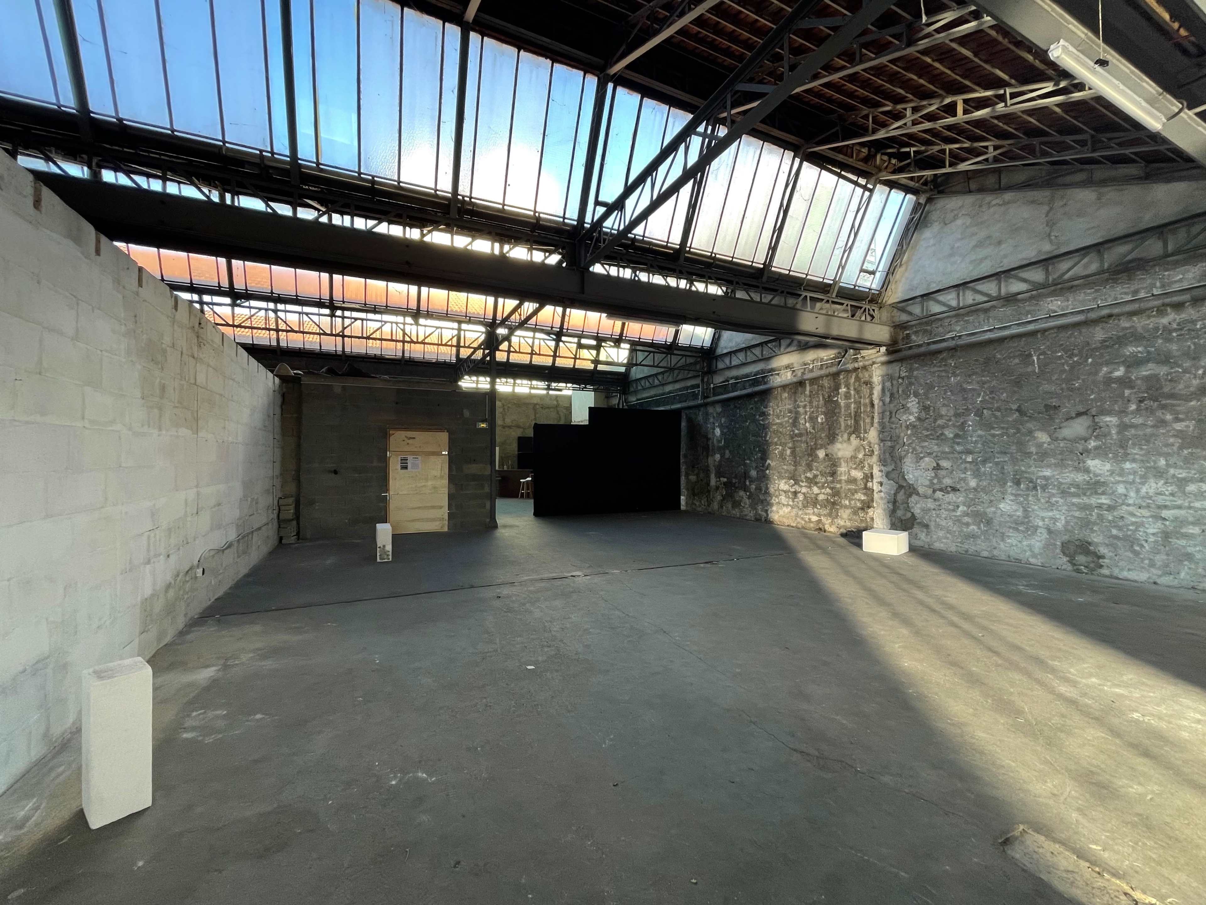 An empty industrial space with concrete walls, large windows, and a blacked-out area in the distance.
