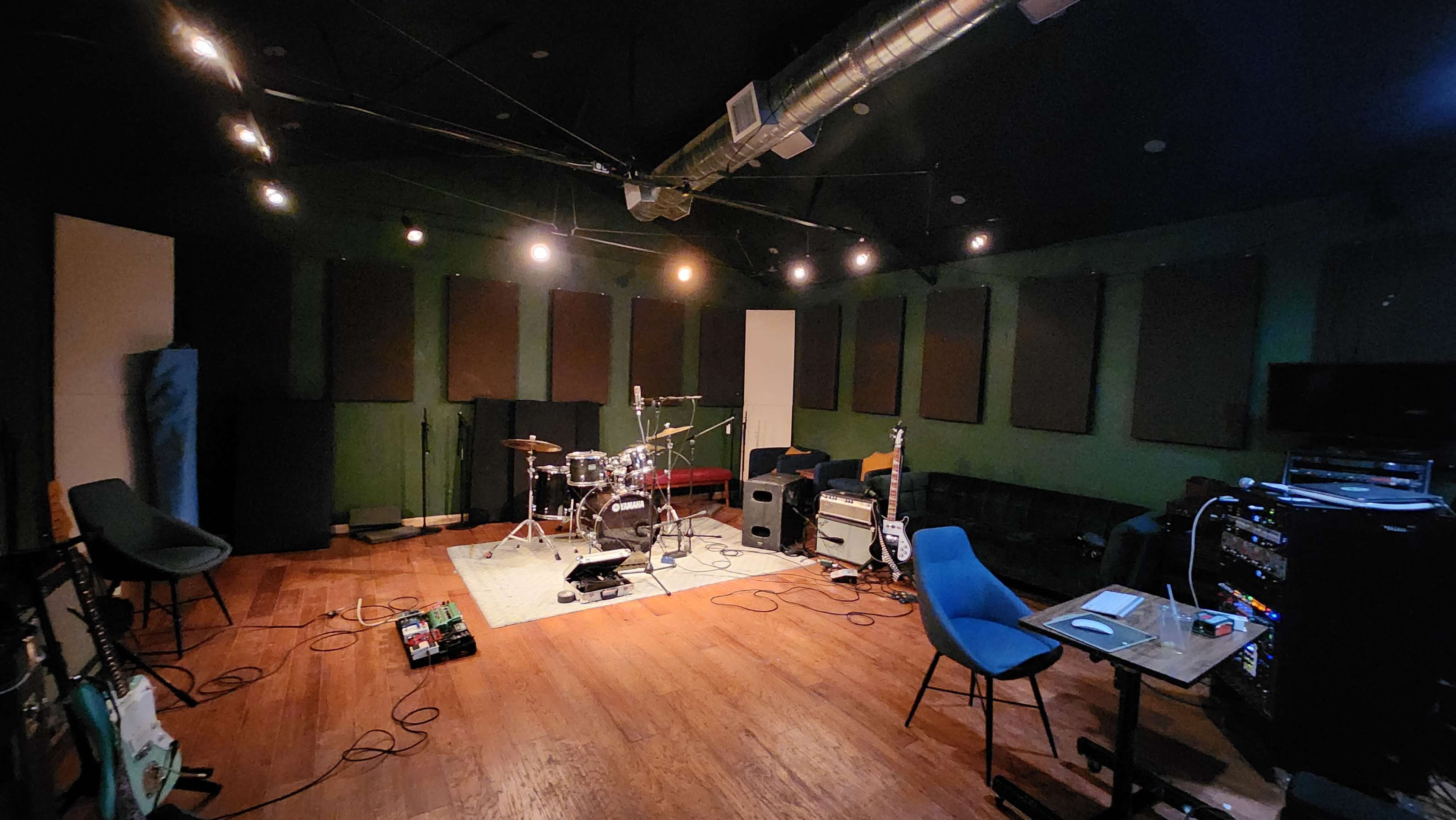 A music rehearsal studio featuring a drum set, amplifiers, and sound equipment arranged on wooden flooring with soundproofing panels on the walls.