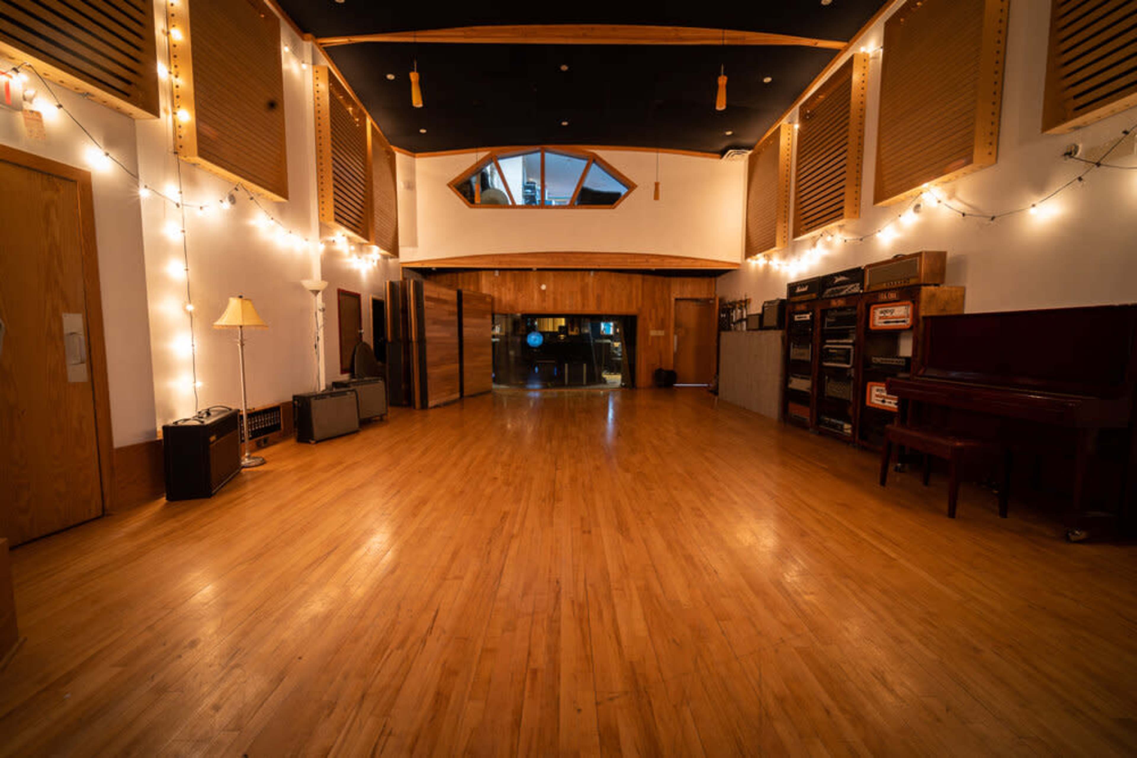 A spacious wooden studio with ambient lighting, amplifiers, and a piano arranged along the walls.