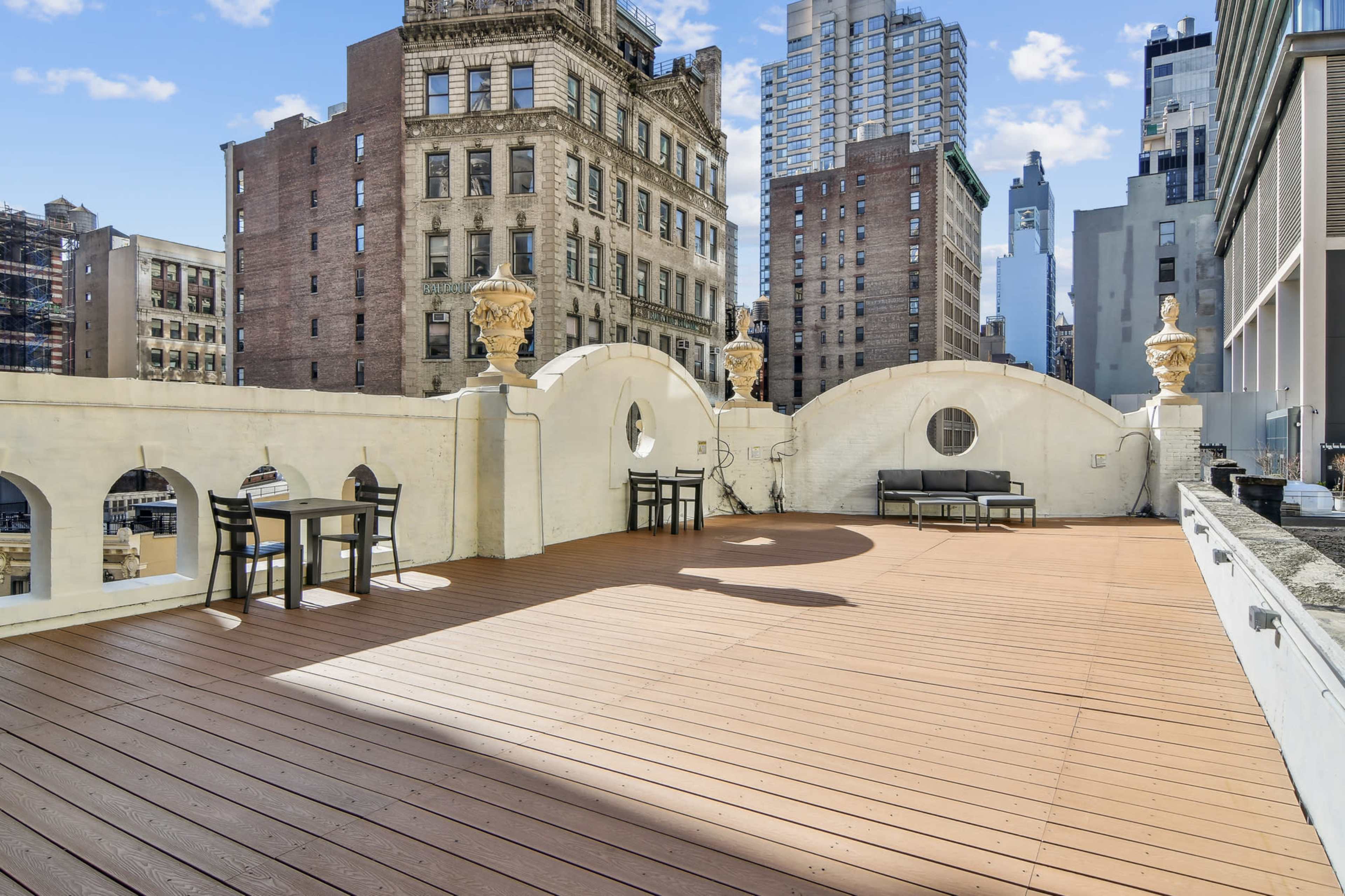 A spacious rooftop terrace with wooden flooring, featuring outdoor furniture and a backdrop of tall city buildings.