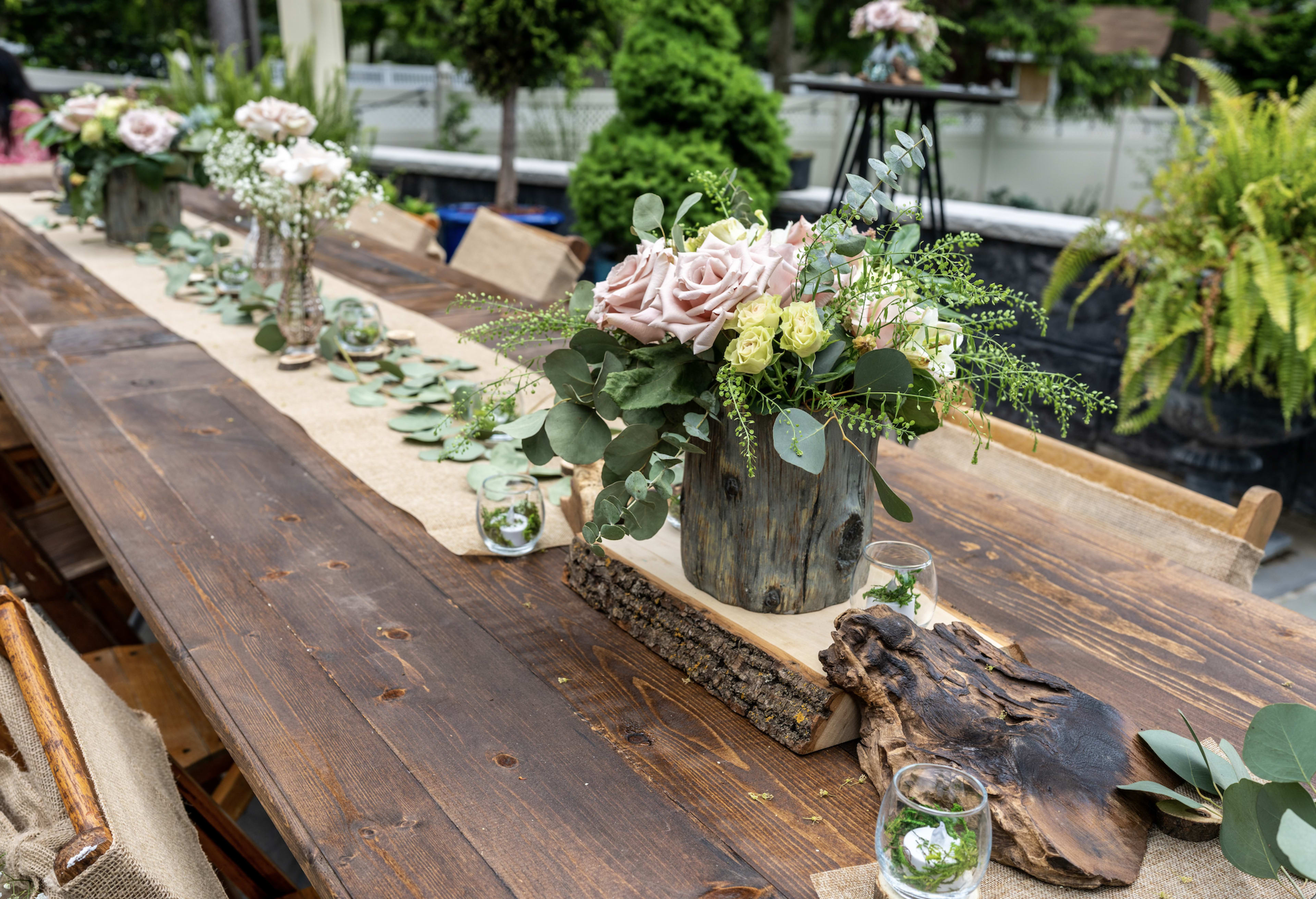 Una mesa de madera con jarrones rústicos llenos de flores en un jardín al aire libre.