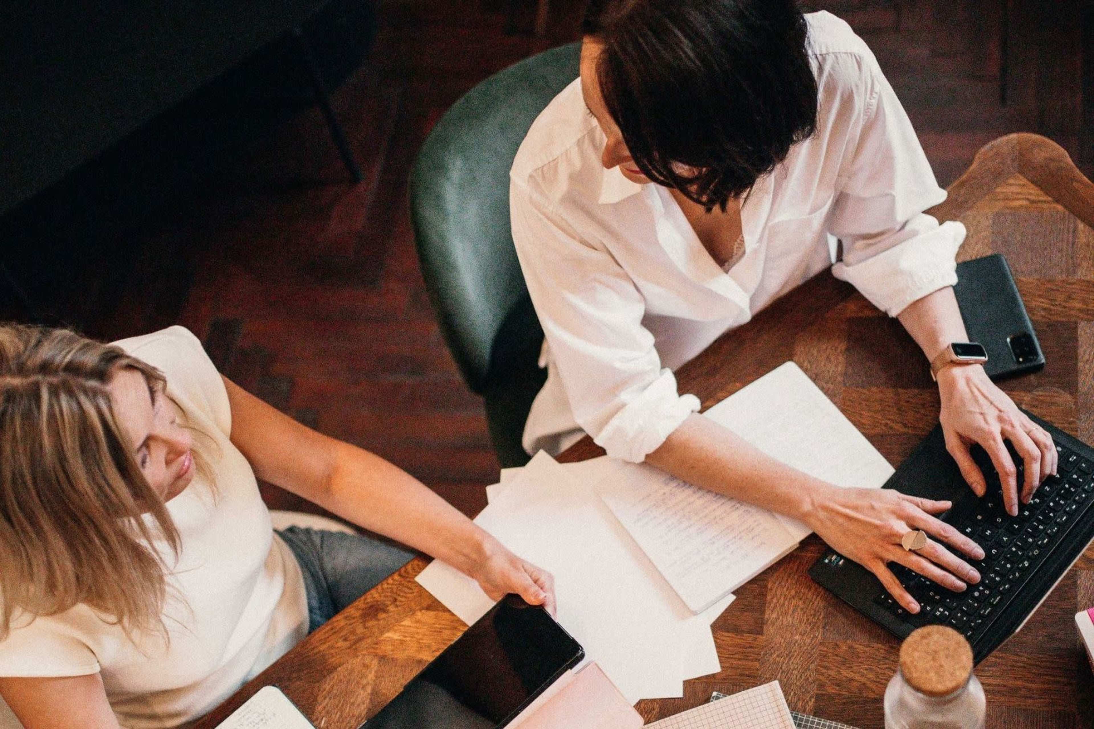 Two women are collaborating at a wooden table, one typing on a laptop while the other looks at a tablet and notes.