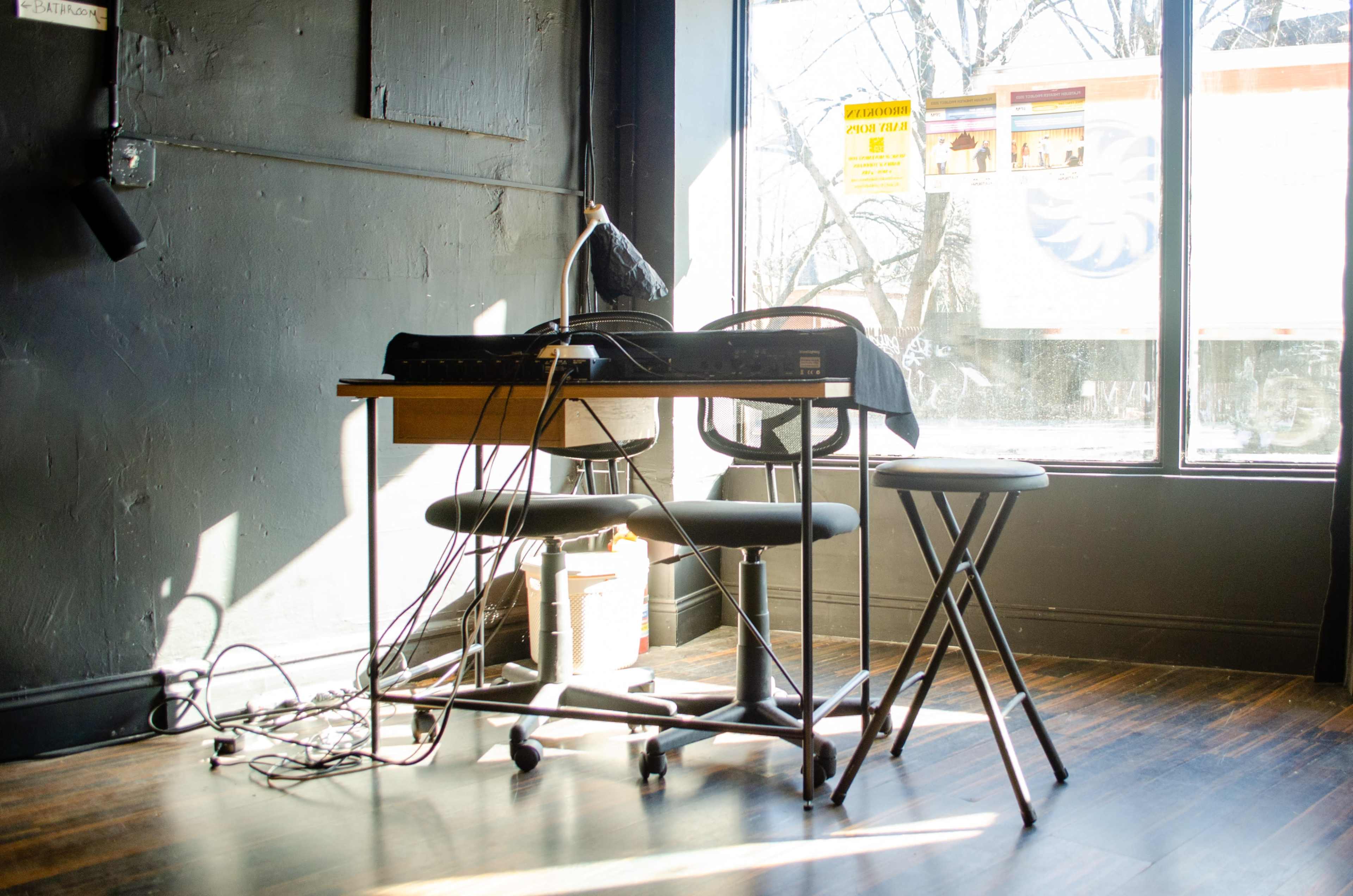 A desk with a black keyboard, a lamp, and a stool is positioned next to a window with sunlight streaming in, revealing tangled cables on the floor.