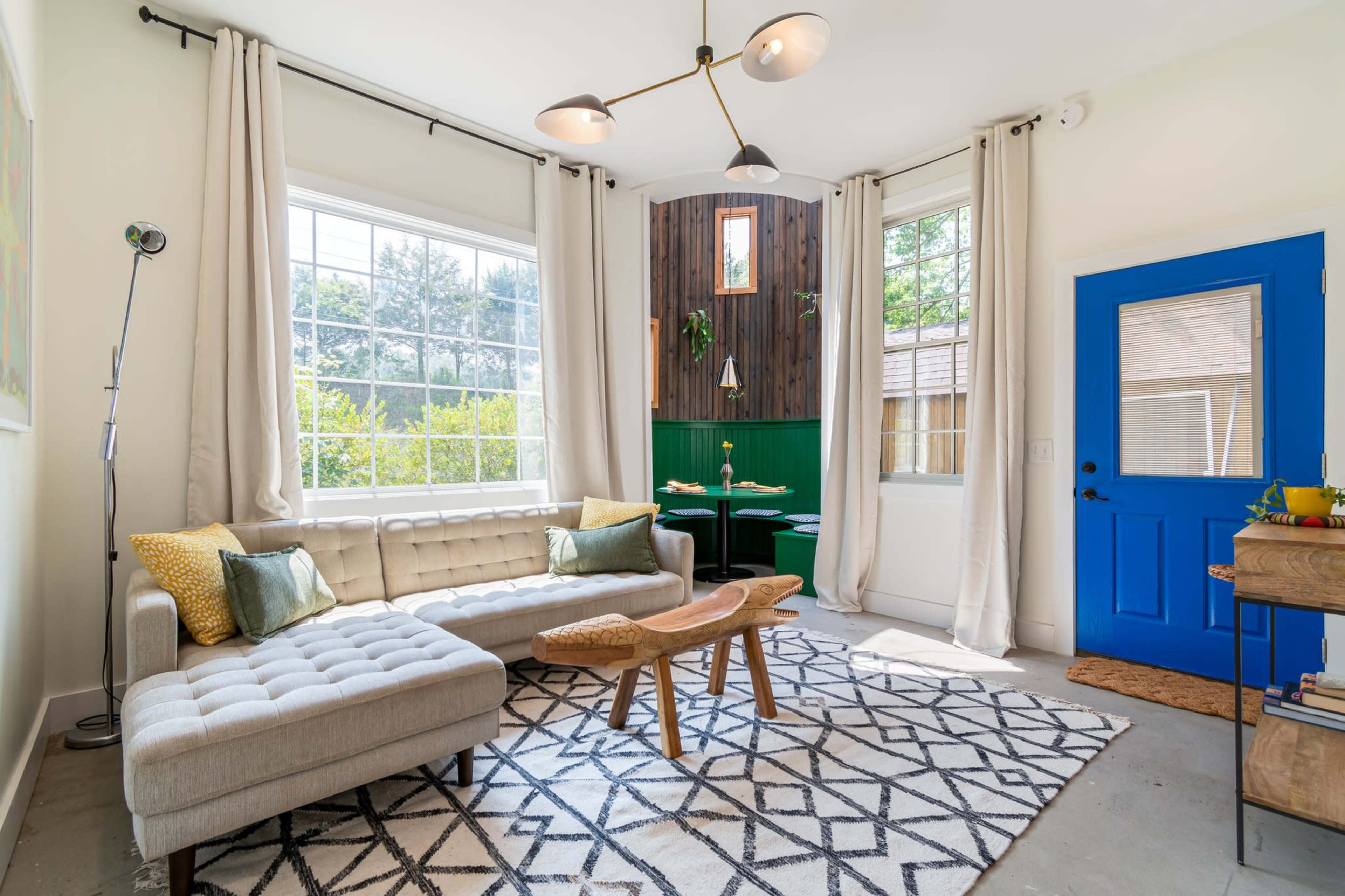 A modern living room features a light-colored sofa, a wooden coffee table, and a bright blue door, with large windows allowing natural light to fill the space.