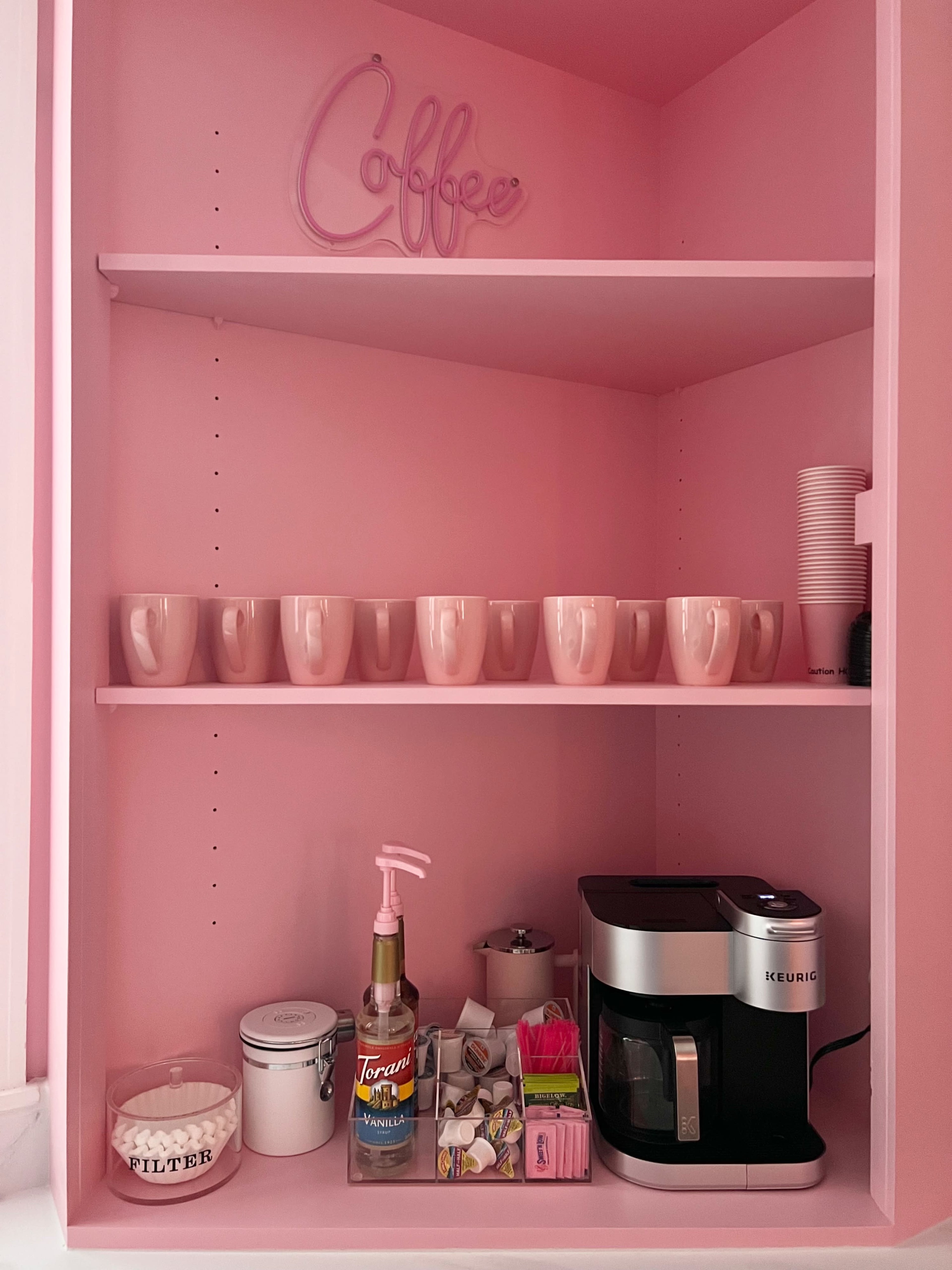 A pink shelf displays a row of pink mugs, a coffee maker, sweeteners, and a container of filters.