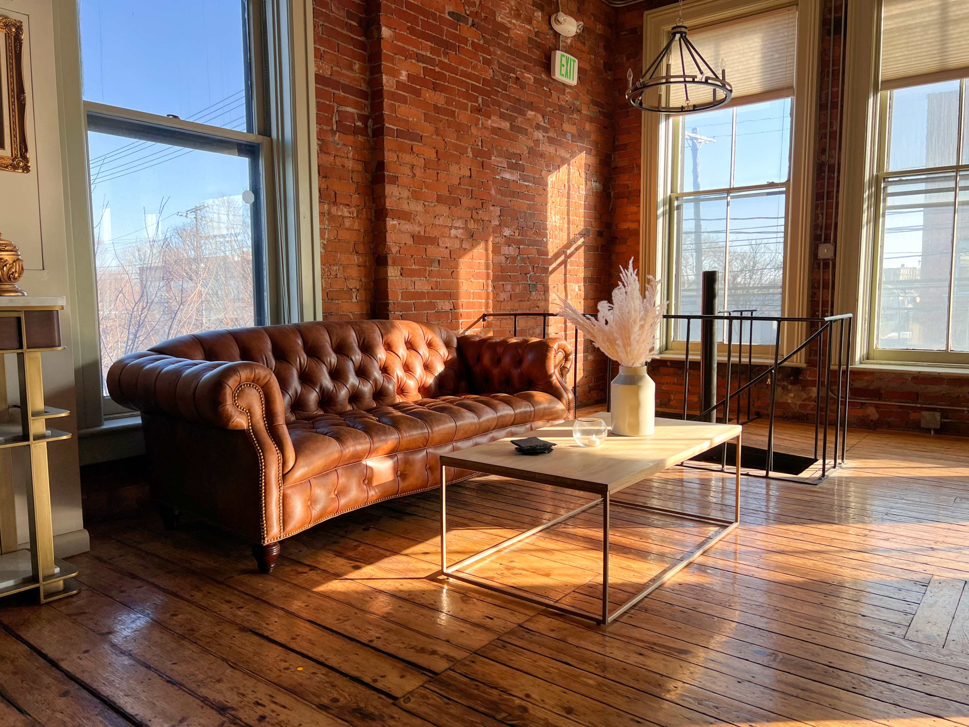A leather chesterfield sofa and a wooden coffee table in a sunlit room with exposed brick walls and large windows.