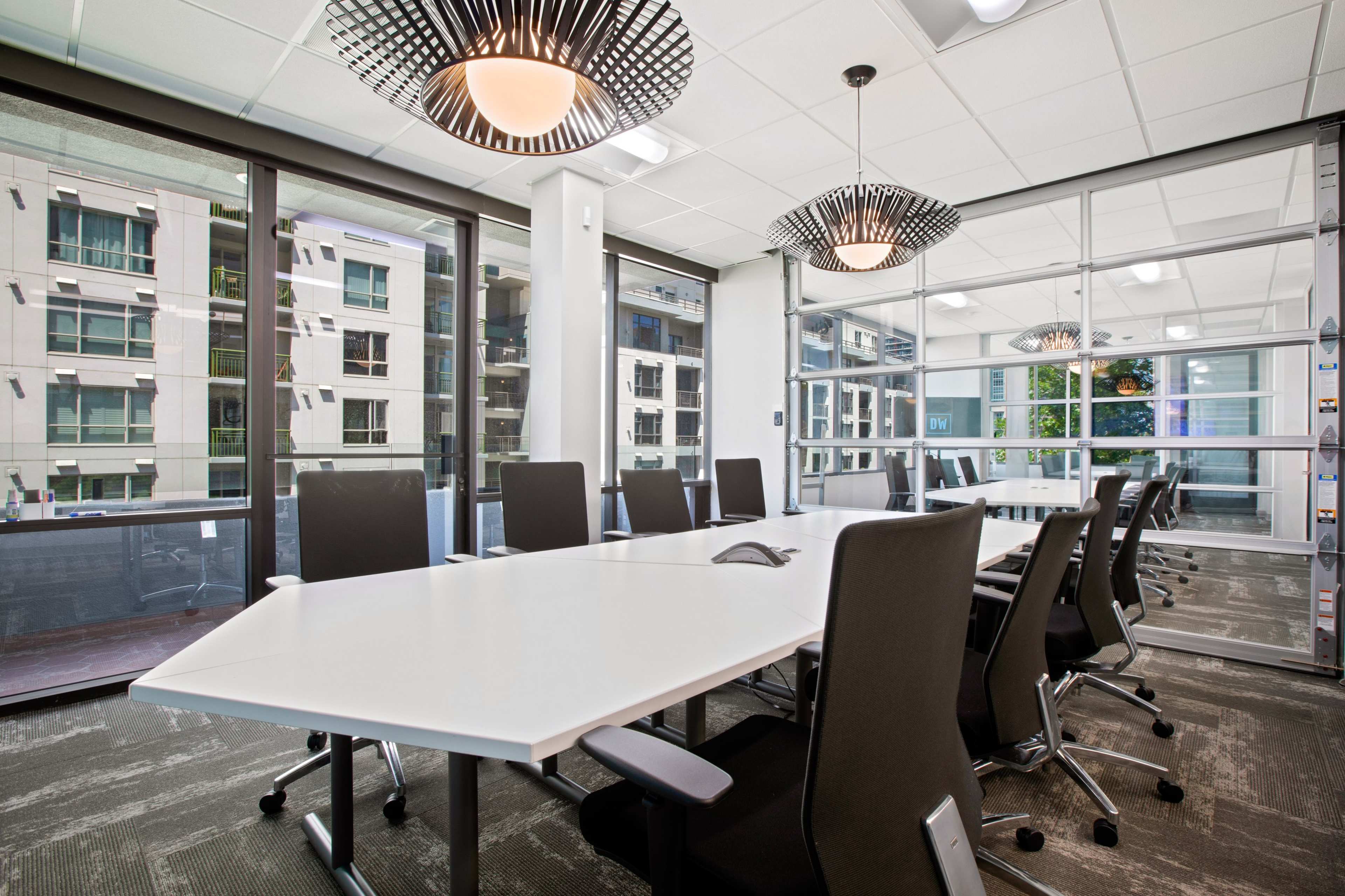 A modern conference room features a large white table surrounded by black rolling chairs and large windows overlooking a building exterior.