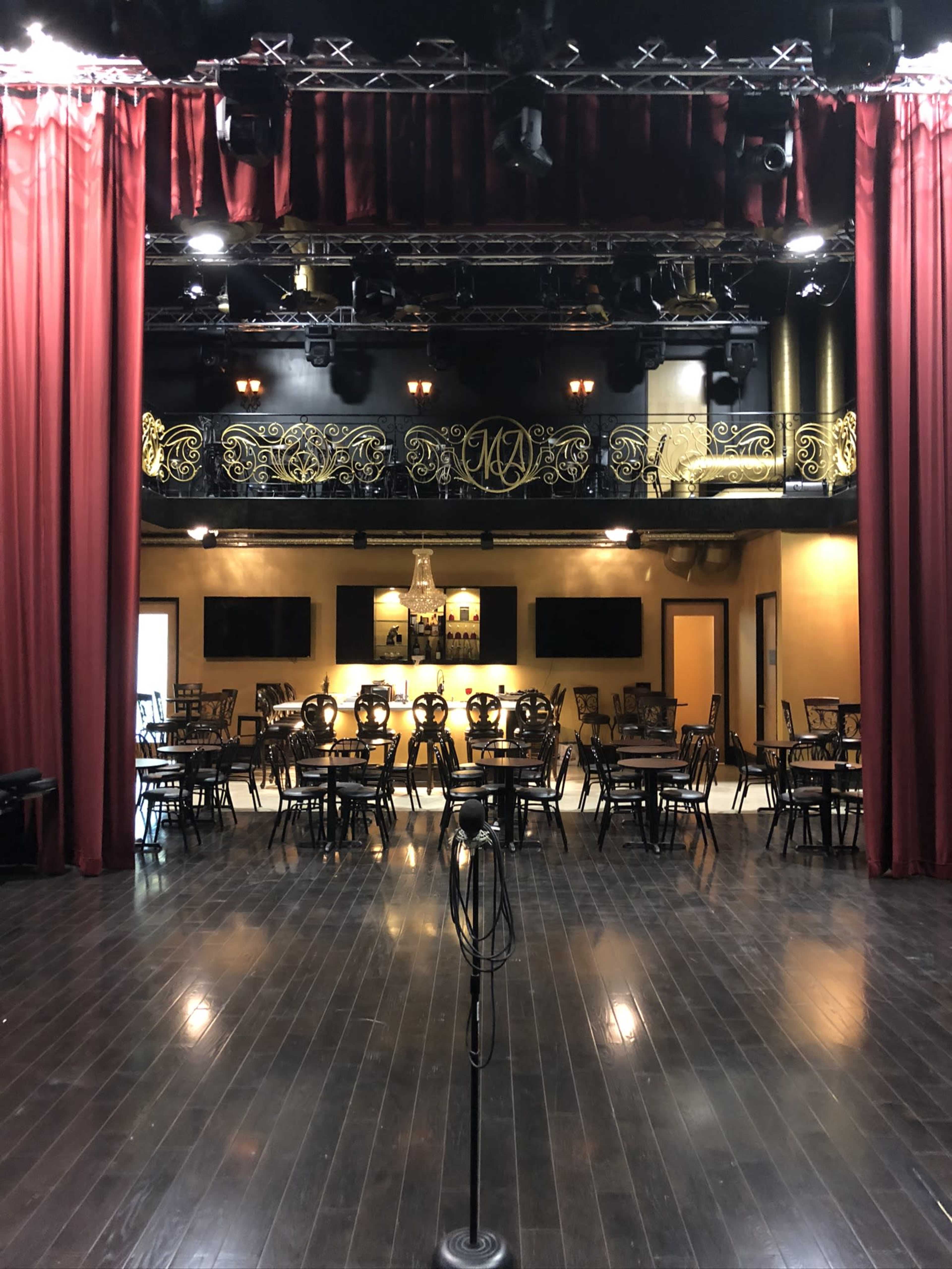 The image shows an empty performance stage framed by red curtains, facing a row of tables and chairs arranged in a dimly lit venue.