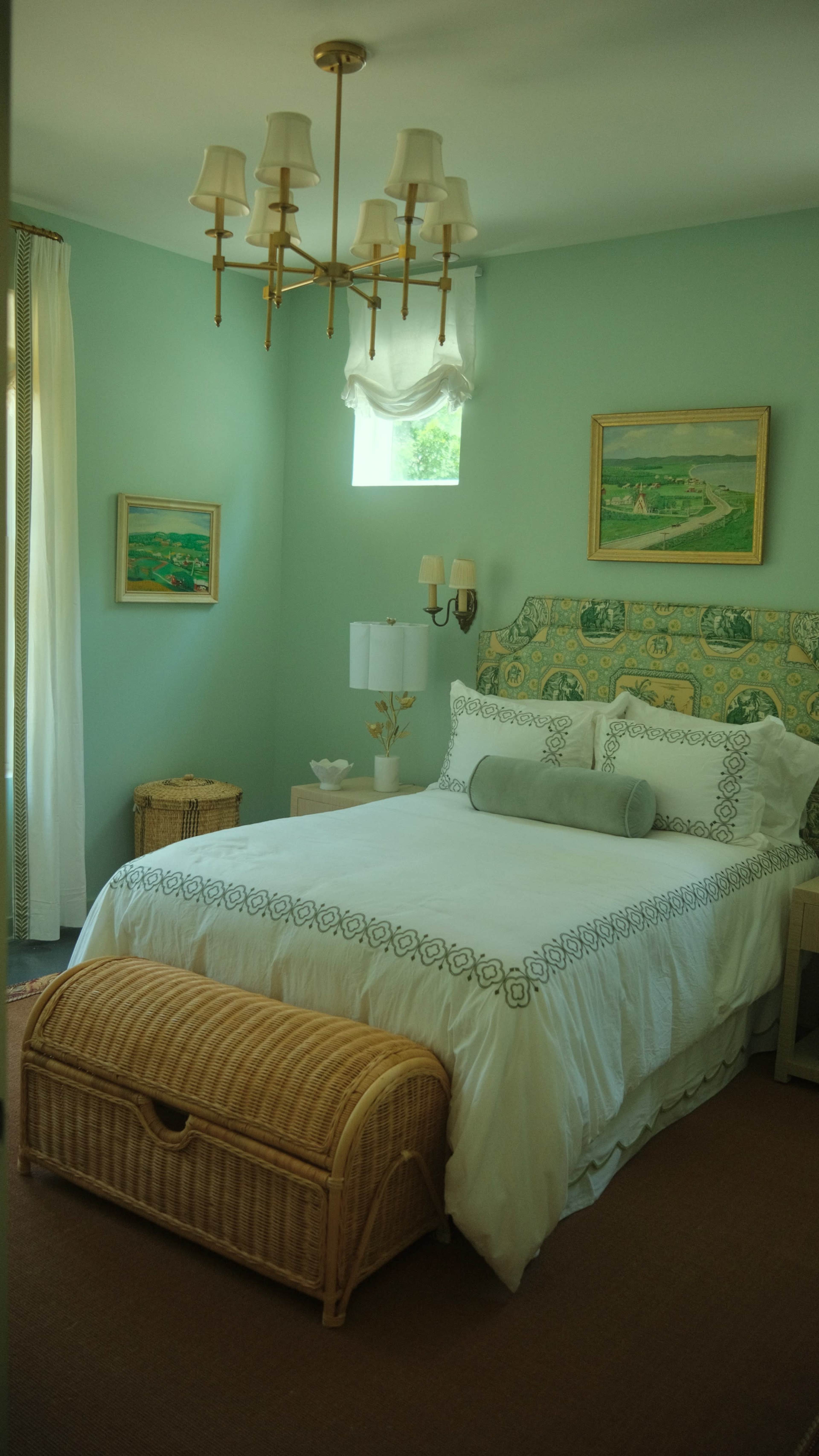 The image shows a bright bedroom with light blue walls, a white bed with decorative pillows, and a woven storage bench at the foot of the bed.