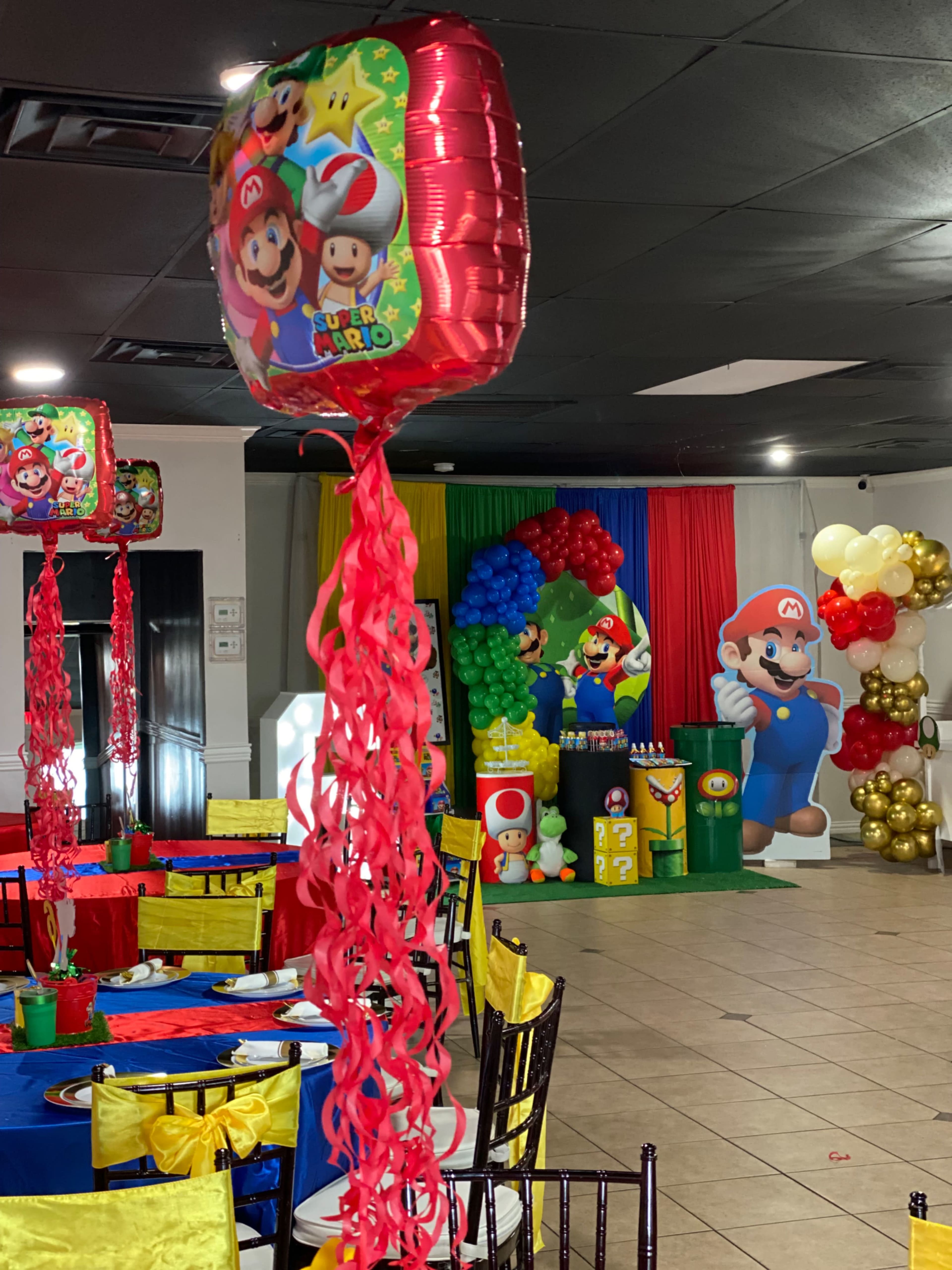 A colorful party room features decorations with a Mario theme, including balloons, themed table settings, and a large backdrop of Mario characters.