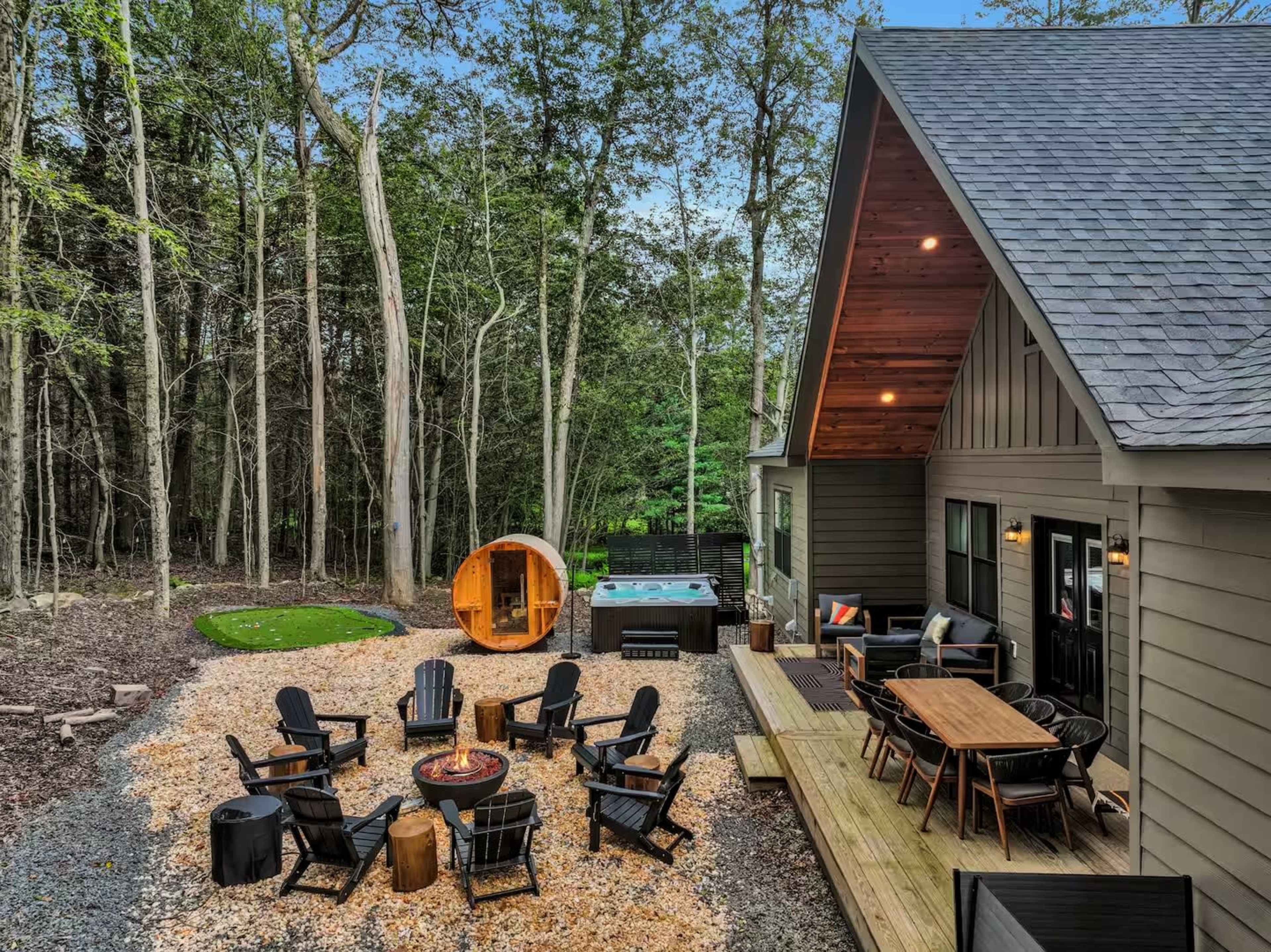 A modern house situated in a wooded area features a patio with a fire pit, seating, and a hot tub nearby.