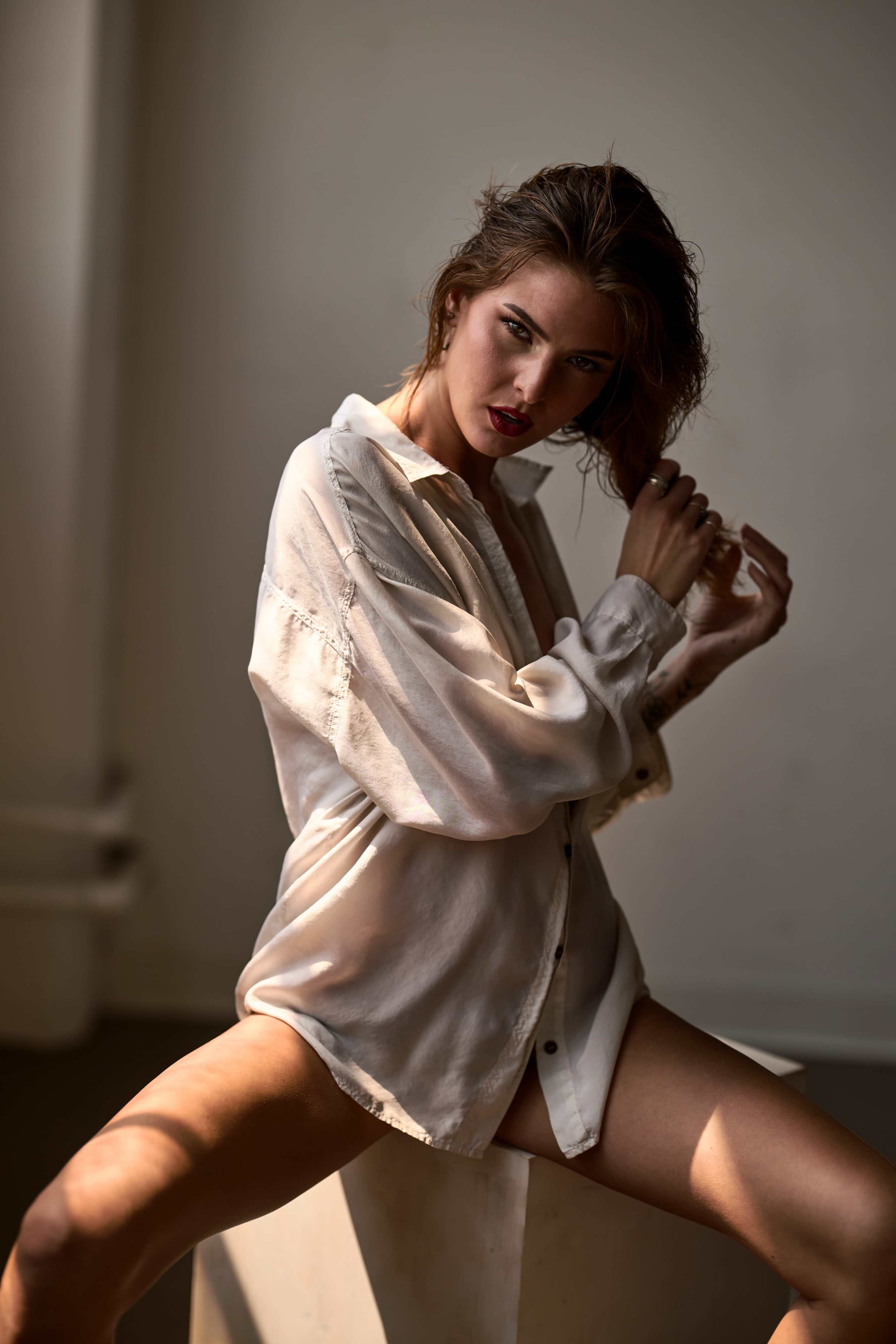 A woman with damp hair poses in a loosely buttoned white shirt, seated on a surface with soft lighting illuminating her figure.