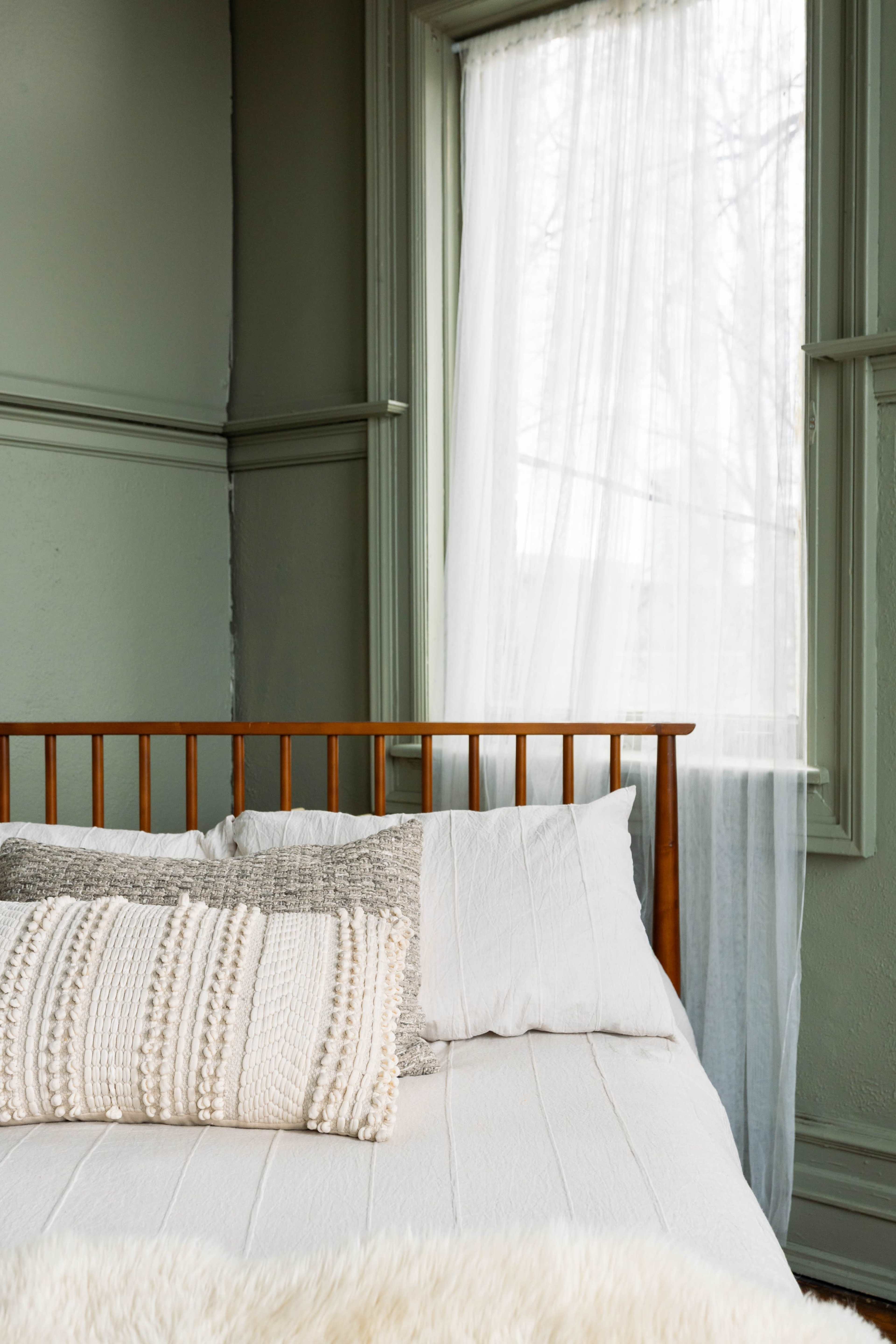 A neatly made bed with decorative pillows sits near a window draped with sheer curtains.
