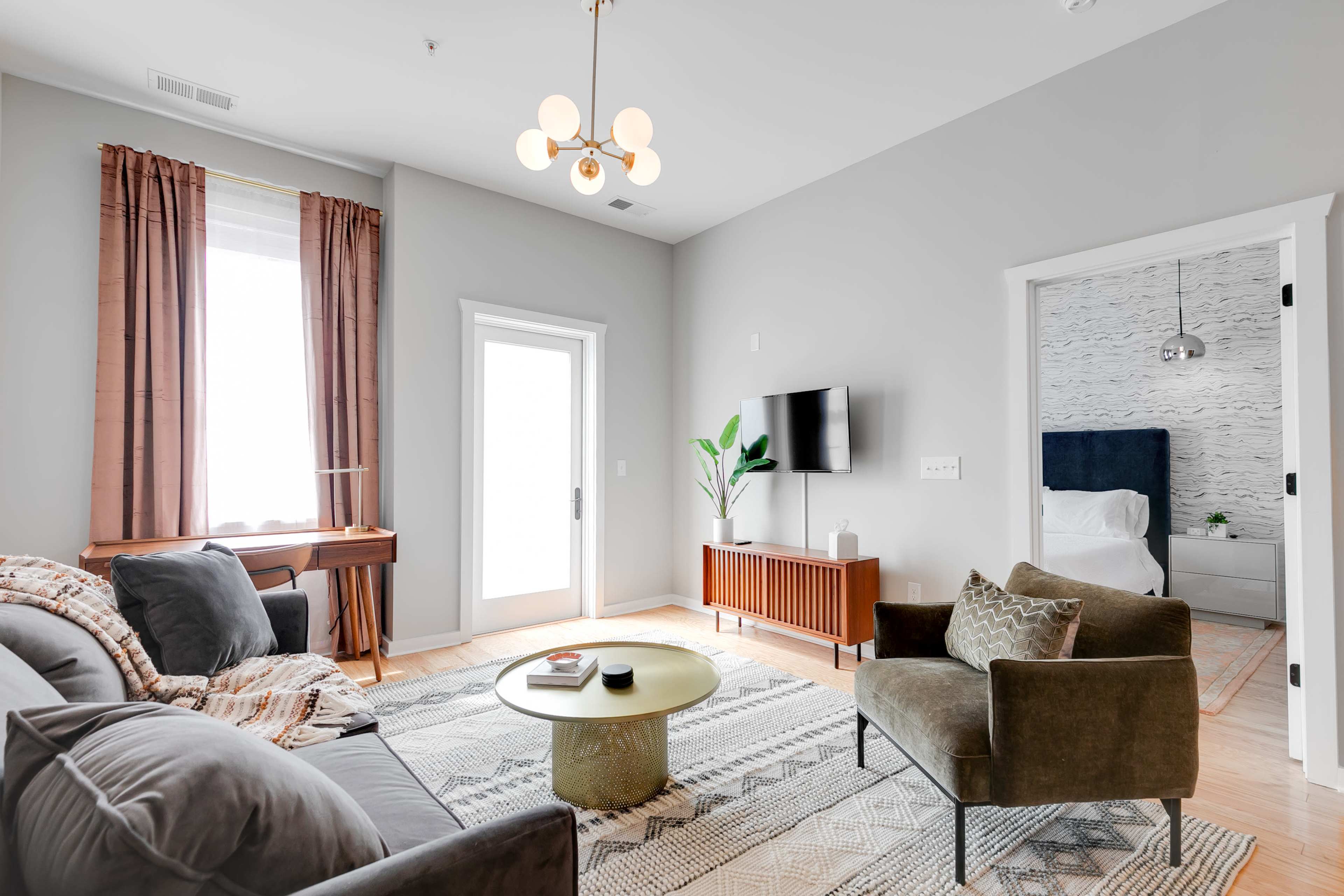 A modern living room features a gray sofa and armchair, a round coffee table, a television mounted on the wall, and large windows with curtains.