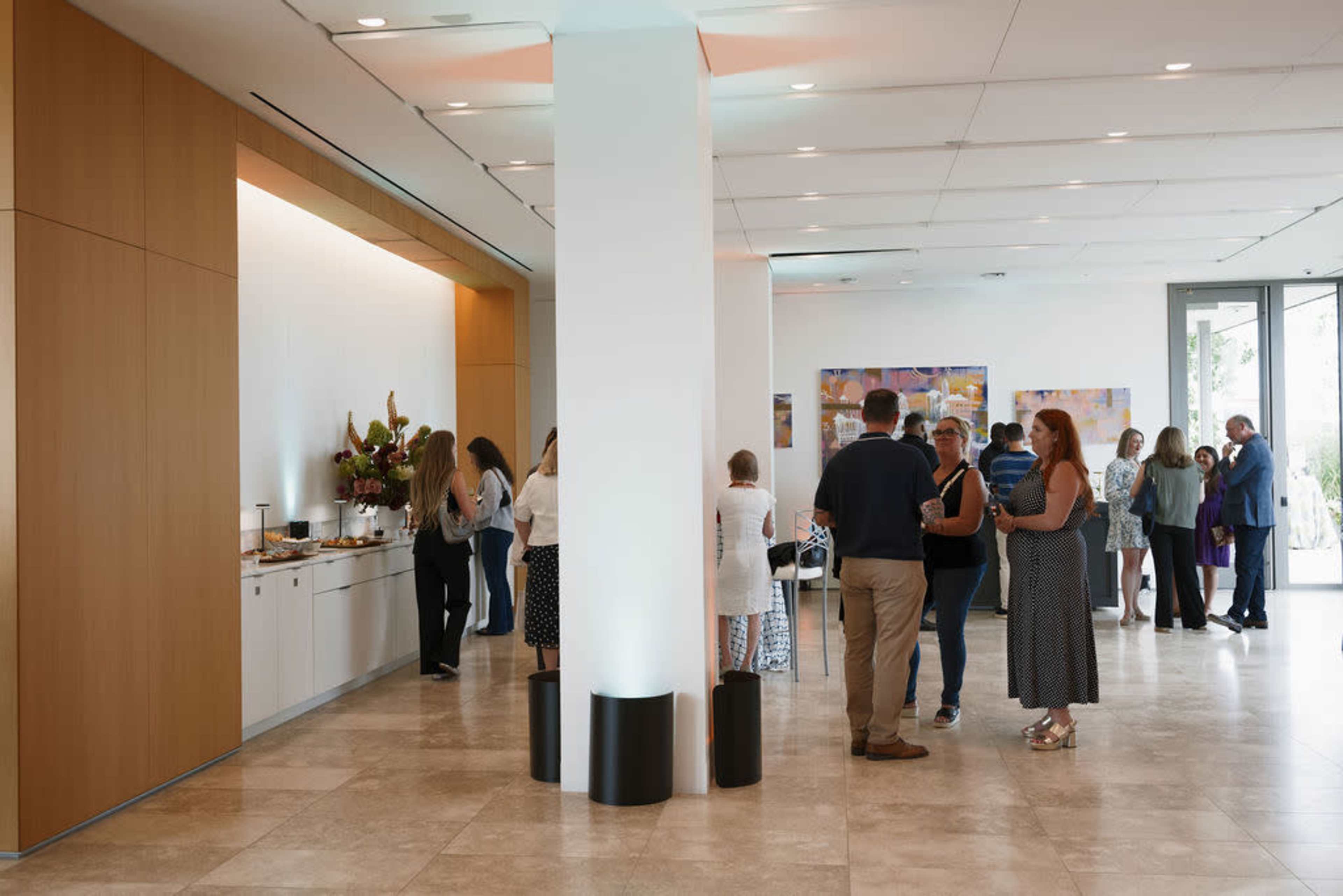 A group of people socialize in a modern indoor space with a snack table along one wall and large windows letting in natural light.