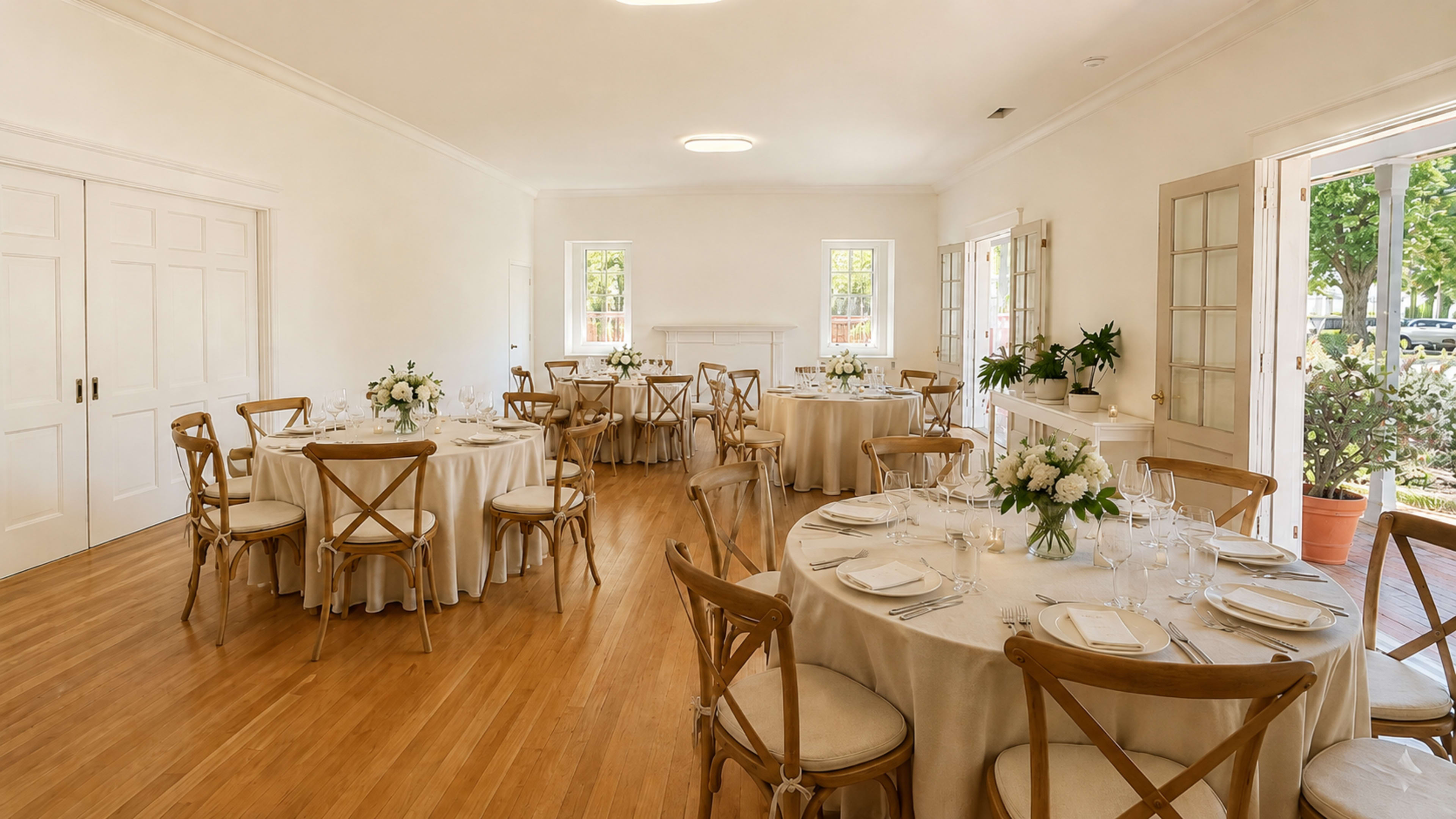 The image shows a bright, elegantly arranged dining room with several round tables set for a meal, adorned with white tablecloths and floral centerpieces.