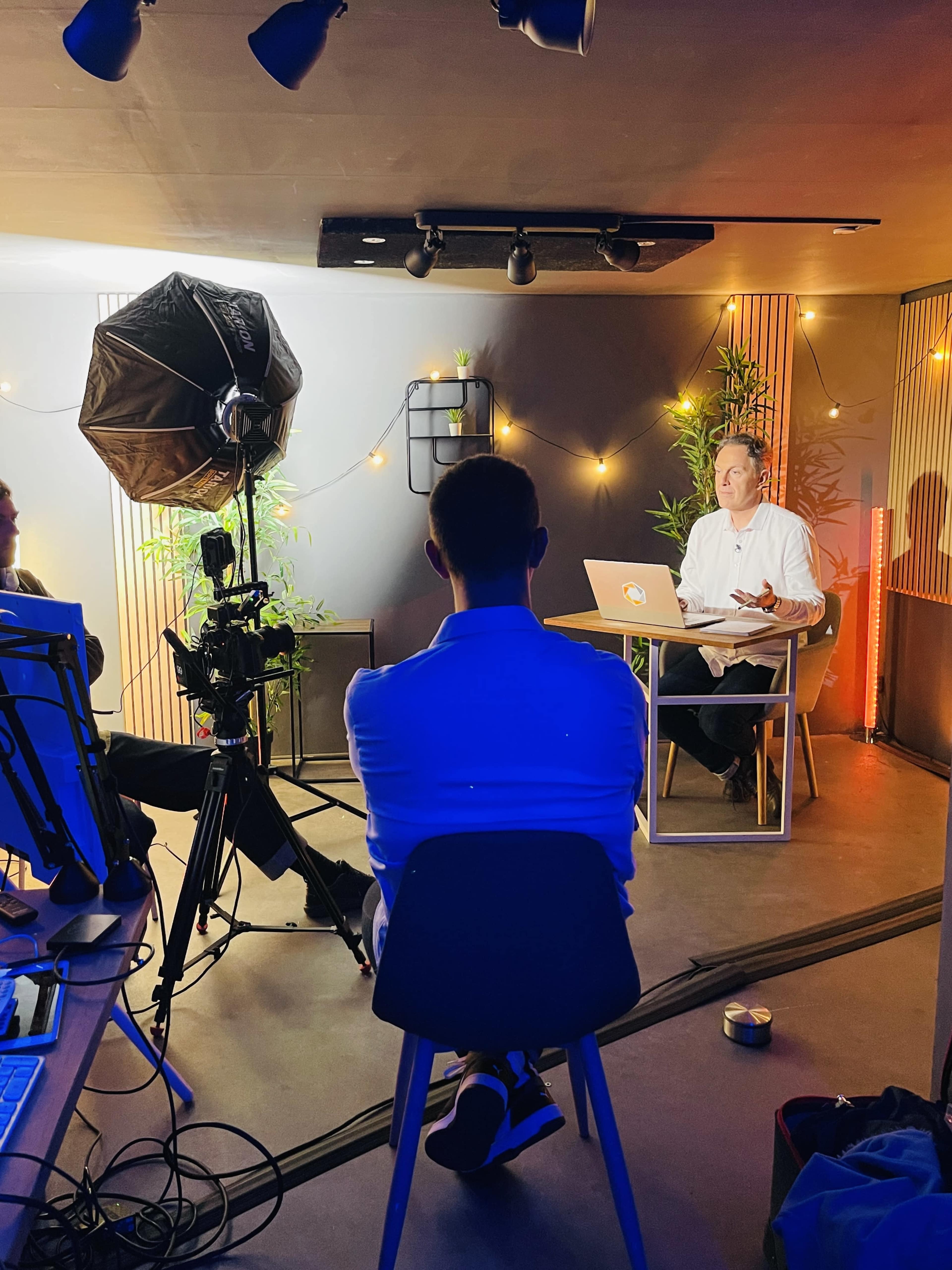 A person in a white shirt is sitting at a table with a laptop while being filmed by a camera in a well-lit studio with decorative plants.