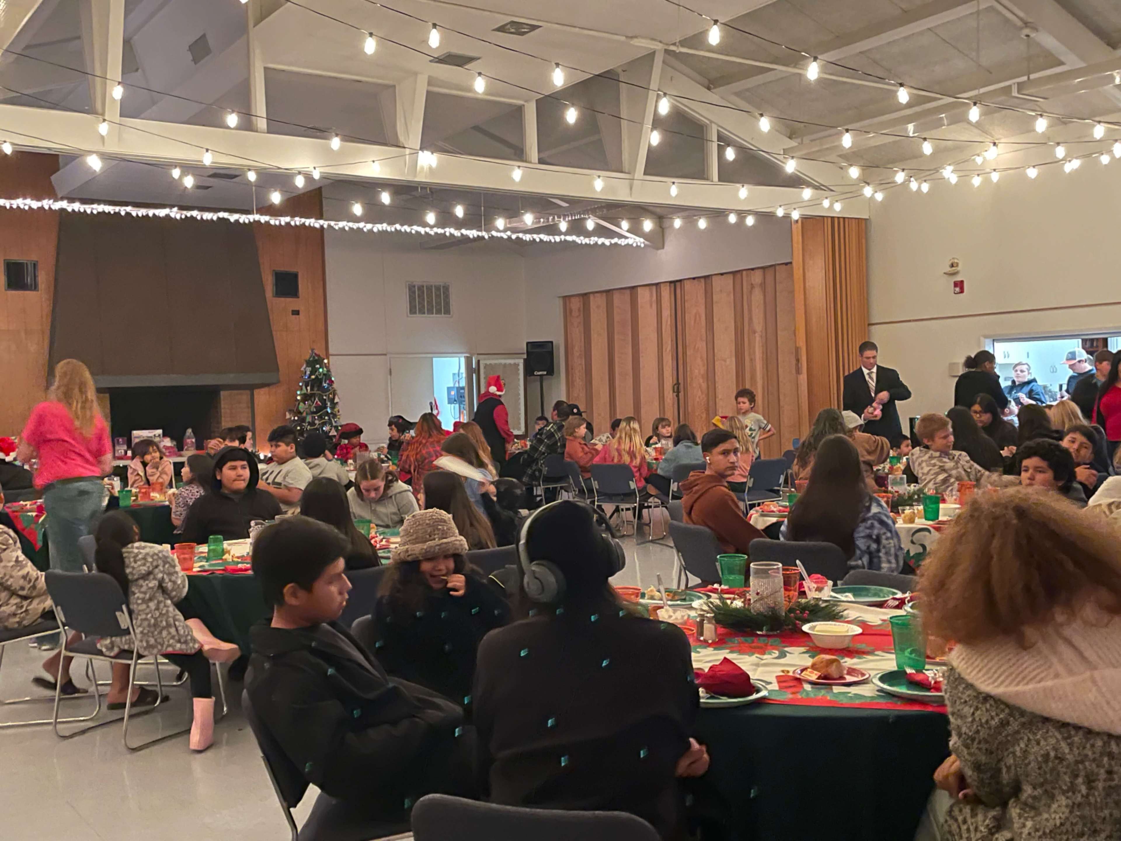 A large gathering in a decorated hall where people are seated at tables set for a festive meal, with holiday decorations and string lights in the background.