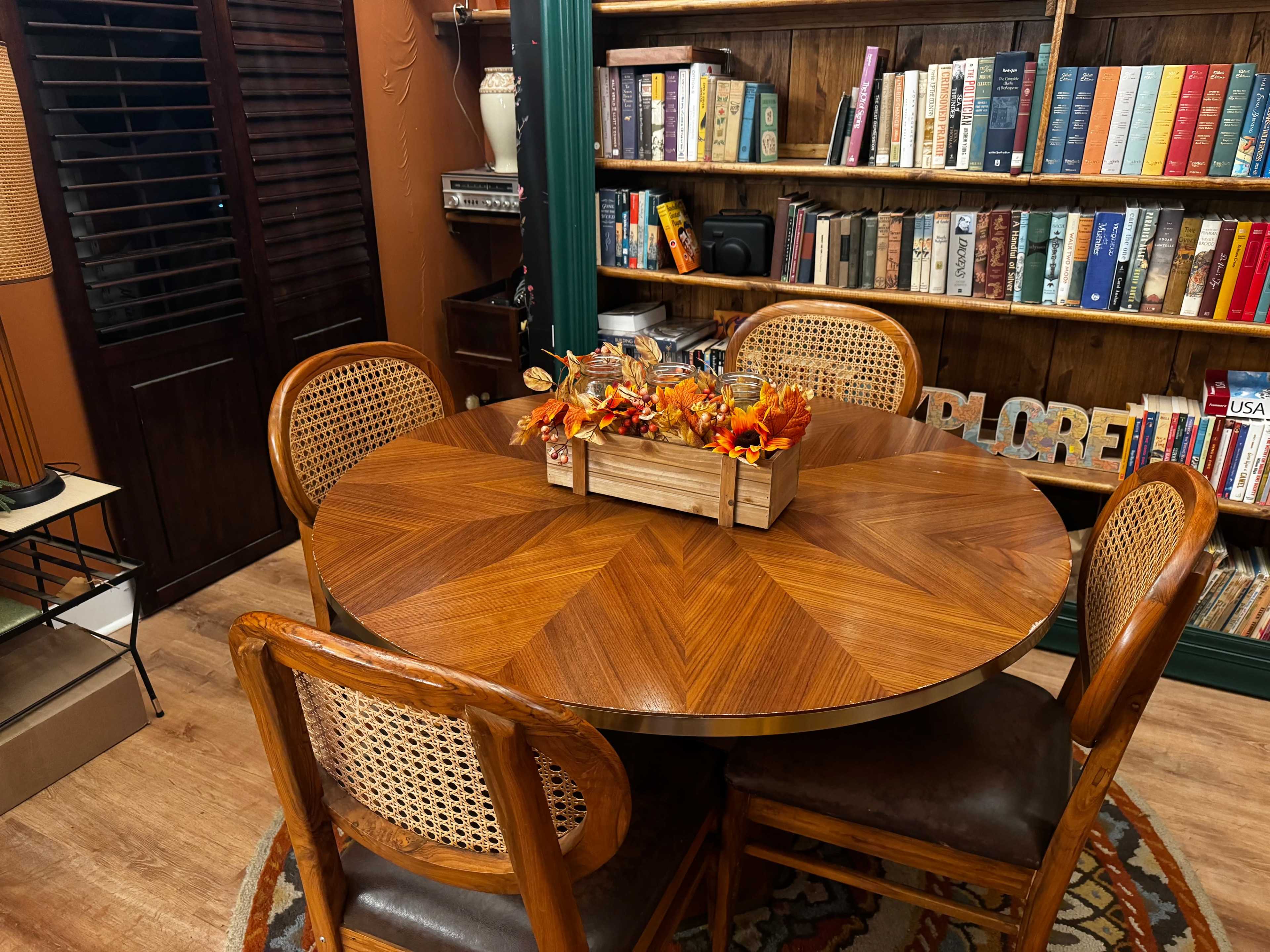 A round wooden dining table is set in a cozy room, surrounded by four cane-backed chairs, with a centerpiece of autumn decorations.